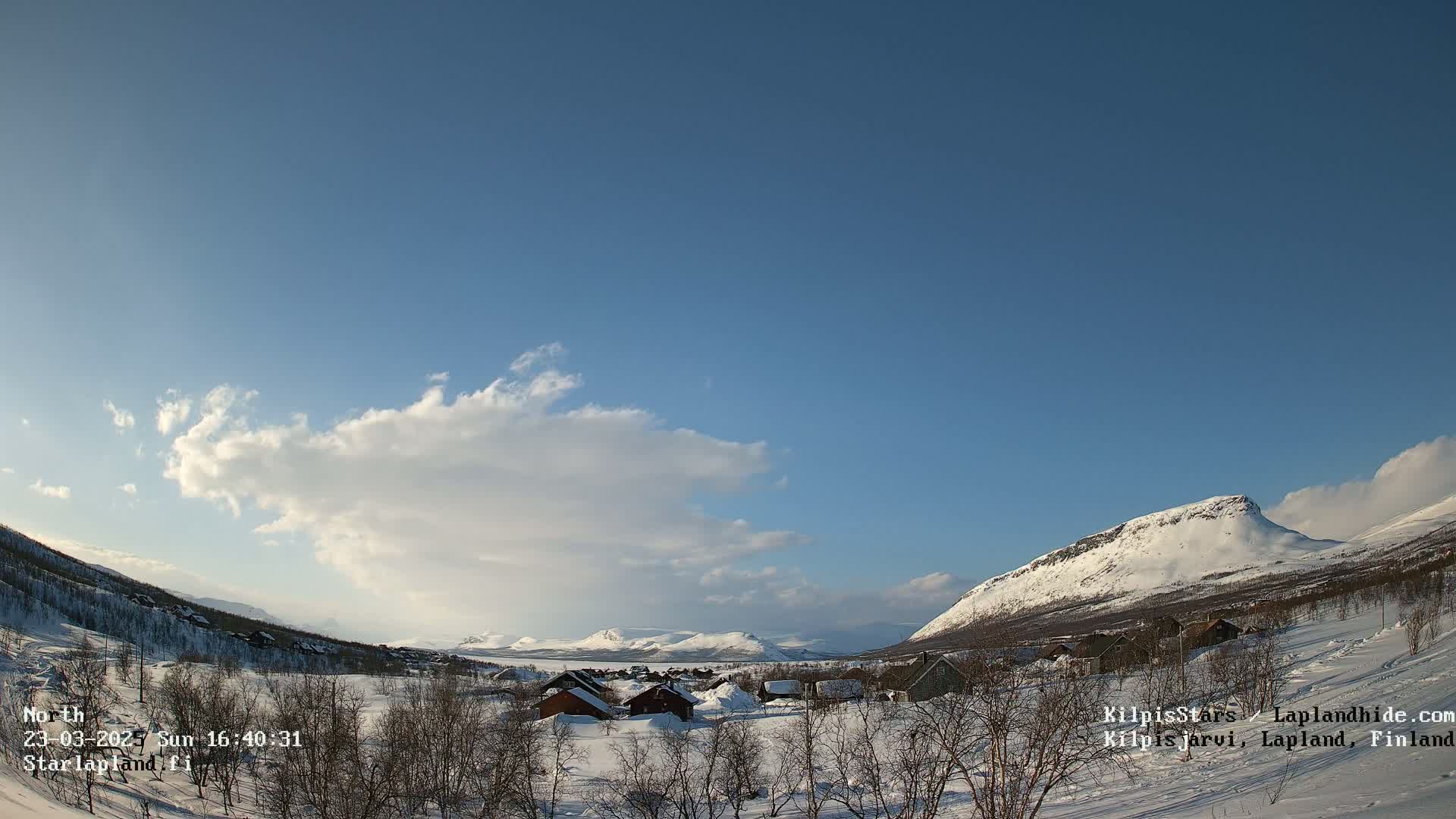 A snow-covered village nestled in a valley between mountains under a partly cloudy blue sky.