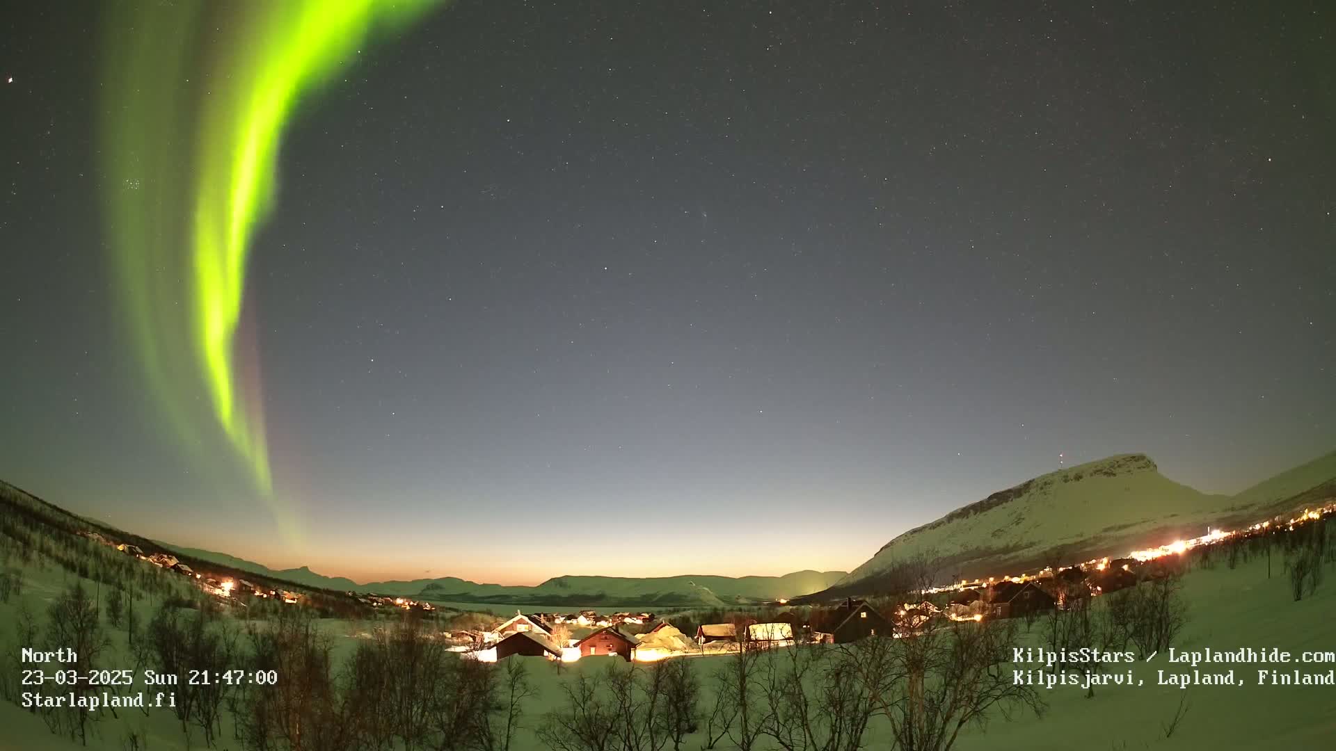 A bright green aurora borealis arches over a snow-covered village nestled in a valley beneath a starry night sky.
