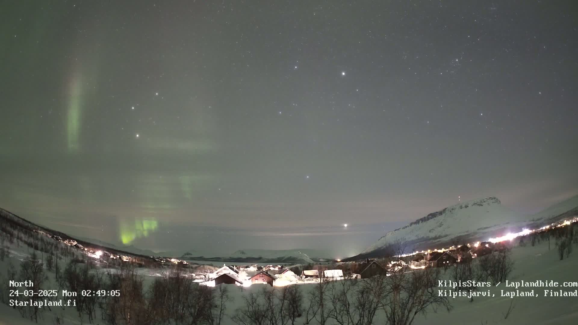 A nighttime view of a snow-covered village nestled between mountains, illuminated by lights, with a vibrant aurora borealis in the sky.