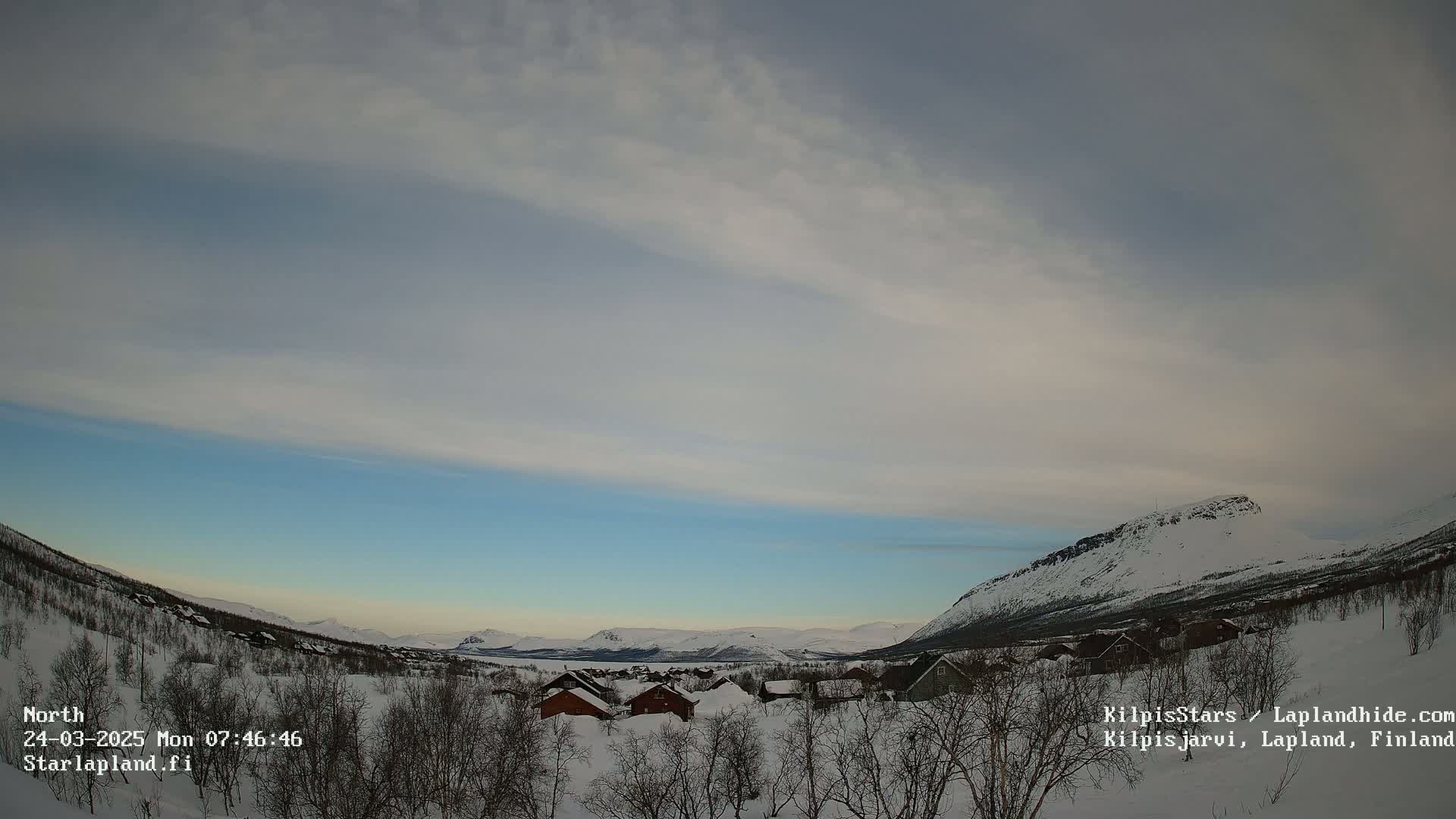 A snow-covered village nestled in a valley between snow-capped mountains under a mostly cloudy sky with a hint of blue near the horizon.