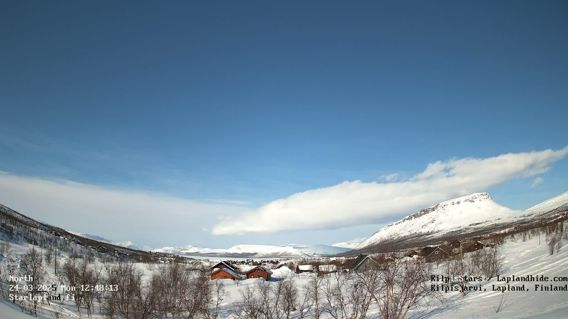 A snow-covered valley with a few small buildings is visible under a mostly clear blue sky with a long, thin cloud formation.