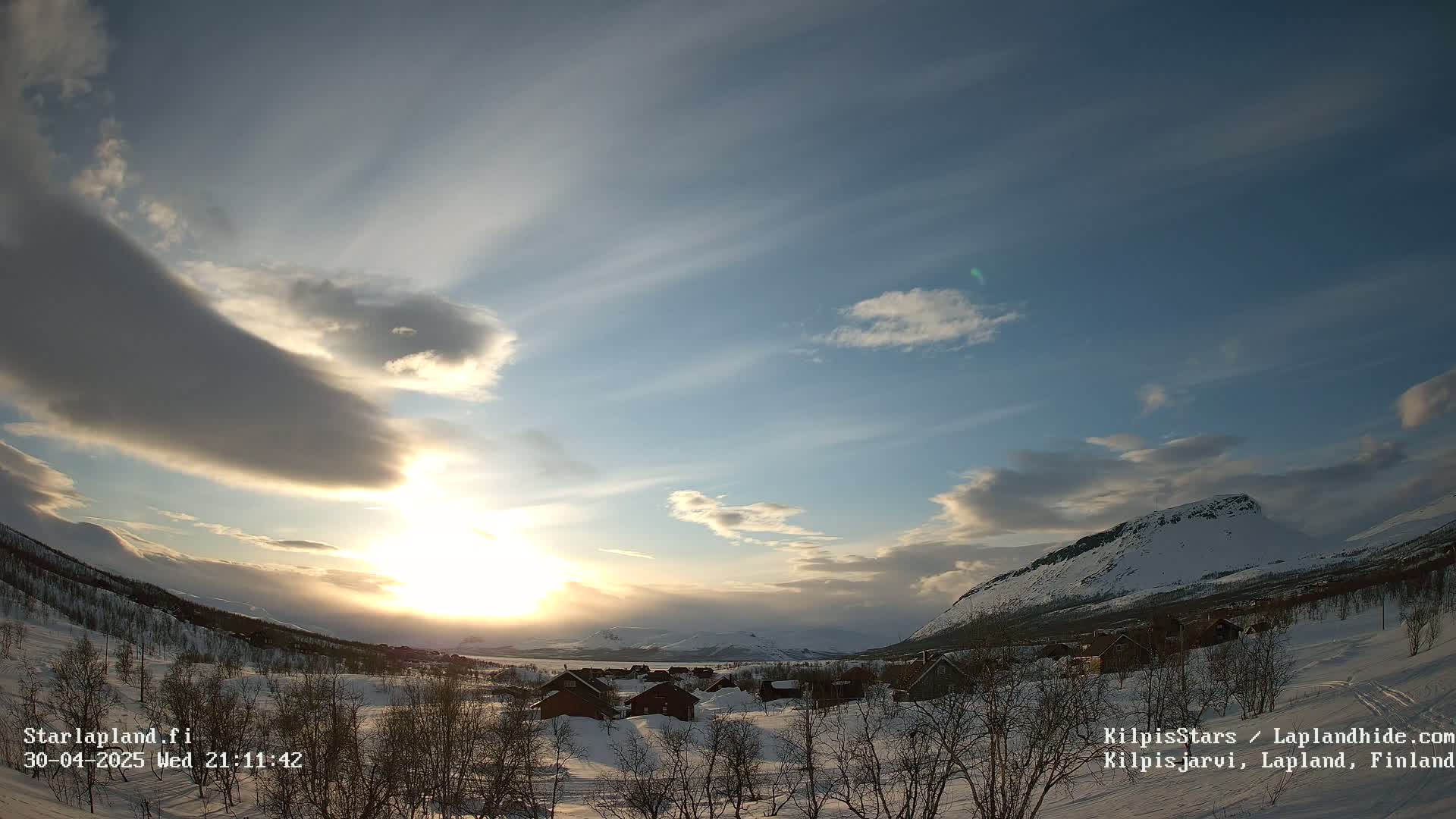 A snowy, mountainous landscape at sunset, with a village nestled in the valley below and a partly cloudy sky.