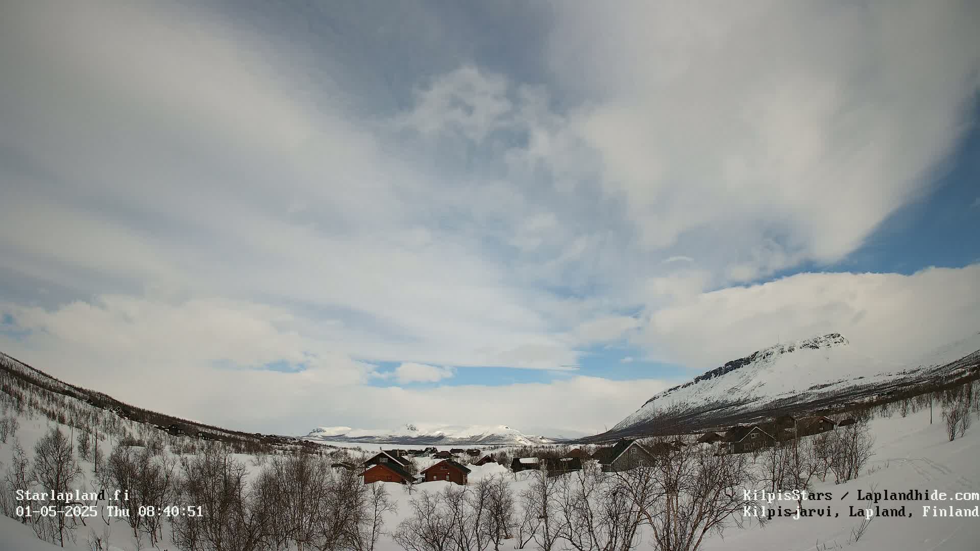A snowy, mountainous landscape with a small village nestled in a valley under a partly cloudy sky.