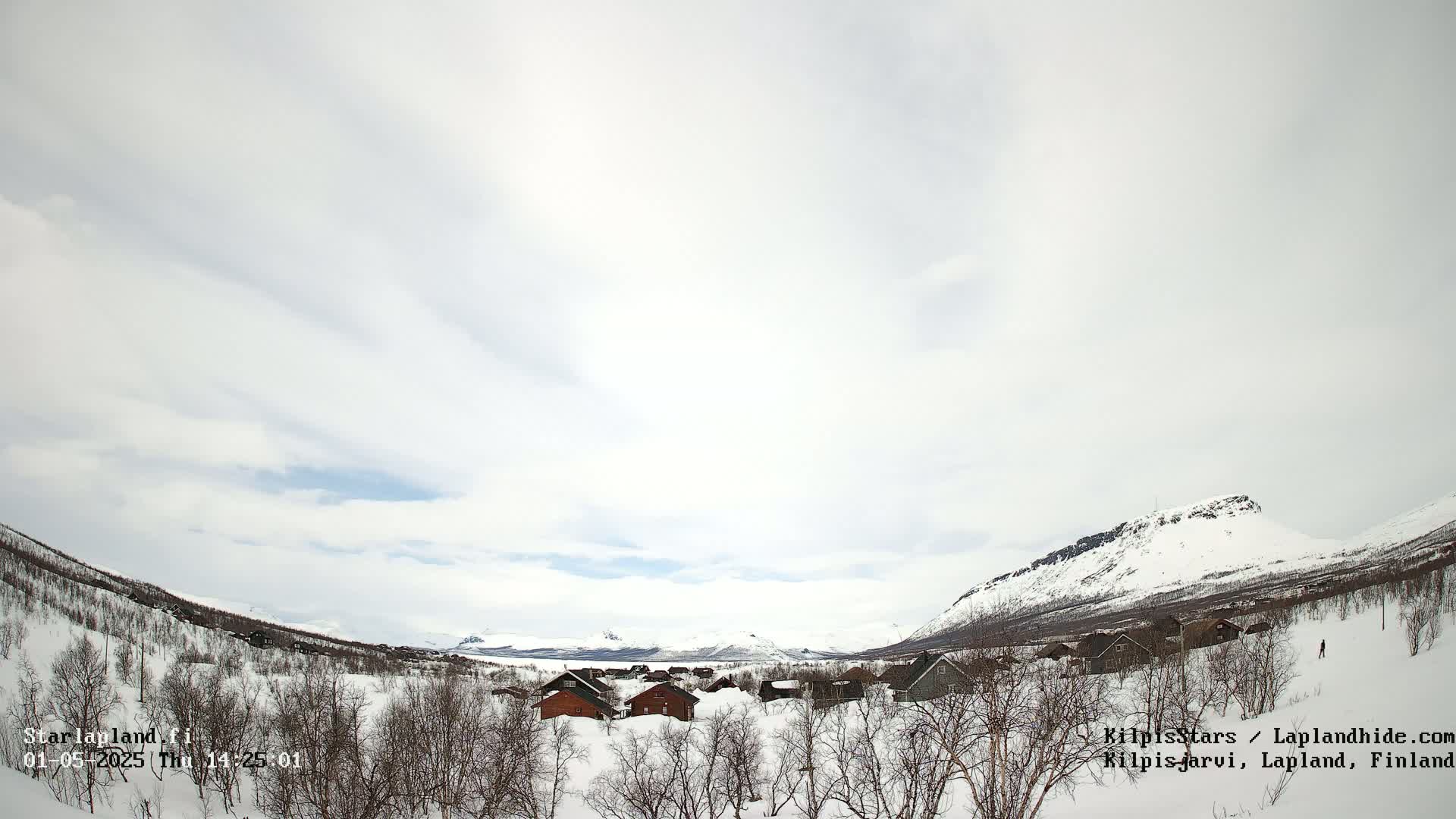 A snow-covered village nestled in a valley between snow-capped mountains under a mostly cloudy sky.