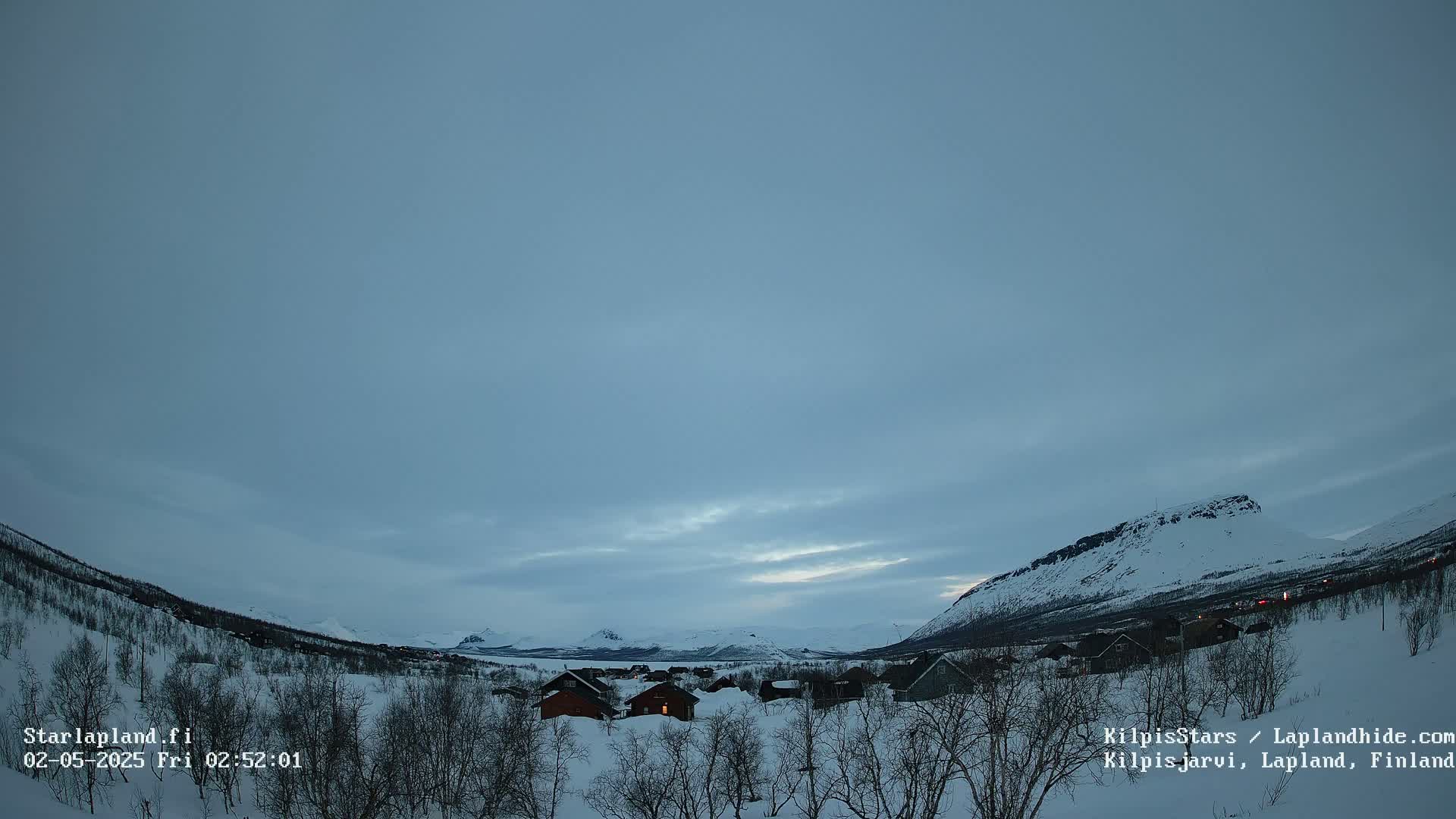 A snow-covered landscape with small houses nestled among leafless trees, mountains in the background under a cloudy, twilight sky.