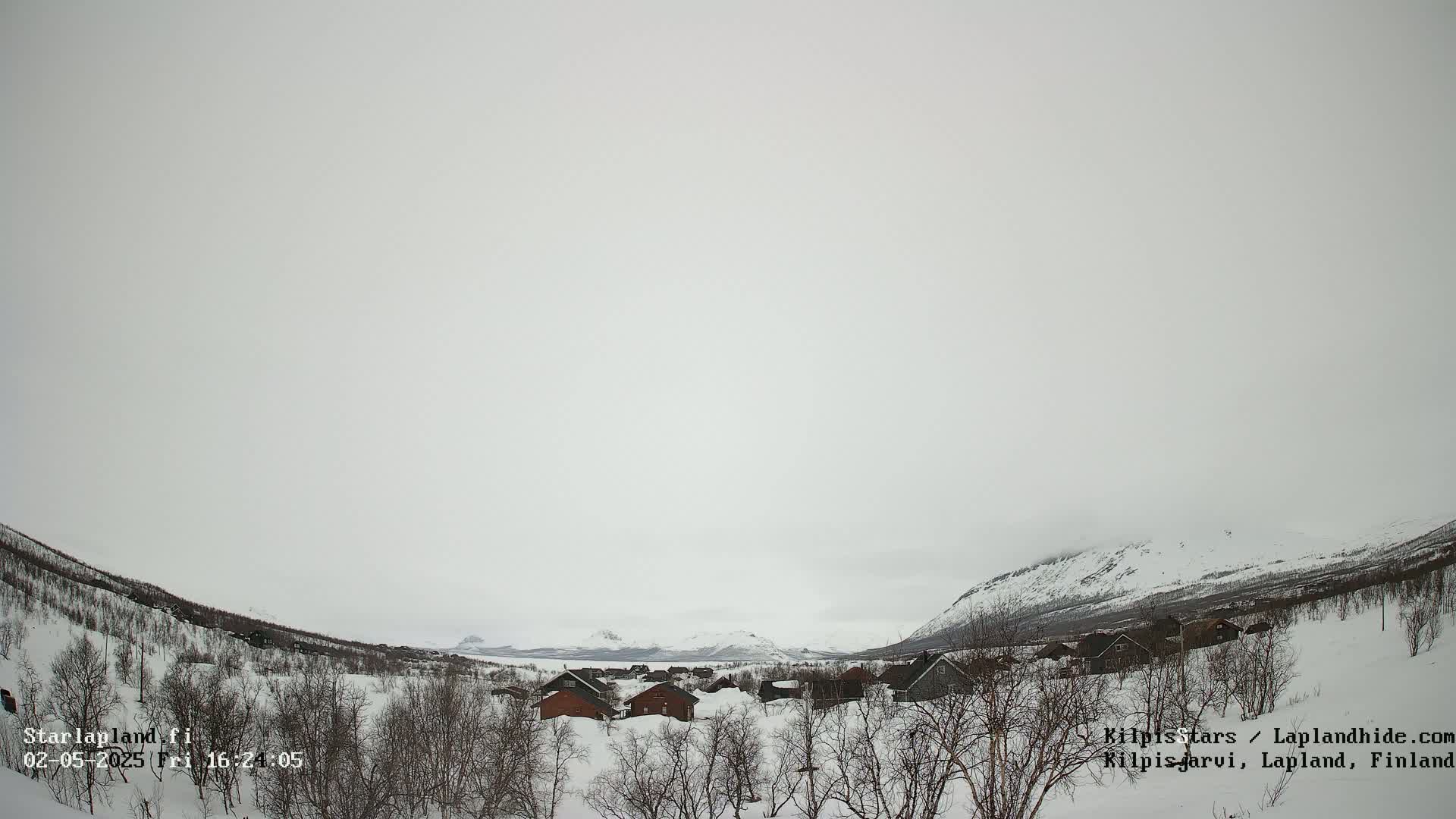 A snowy, overcast landscape shows a small village nestled between snow-covered hills.