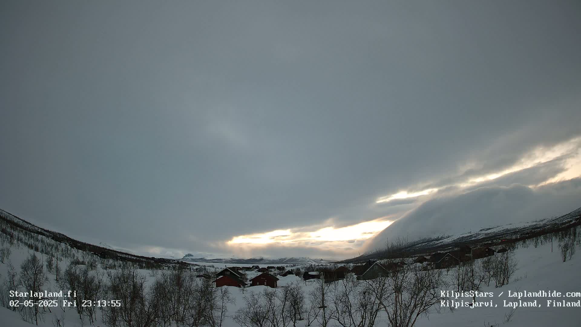 A snowy, winter landscape with a small village nestled among snow-covered hills, under a mostly cloudy sky with a sliver of sunlight breaking through.