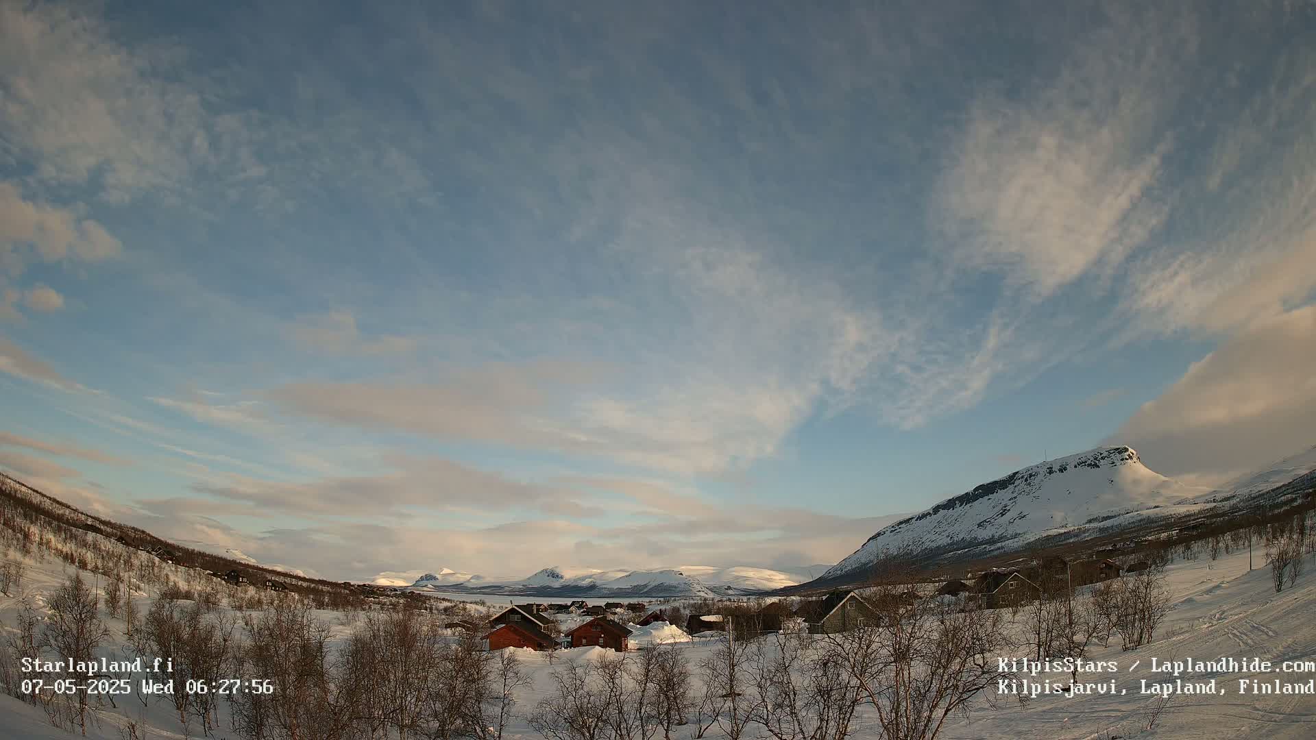 A snow-covered village nestled in a valley between mountains under a mostly clear sky with wispy clouds.