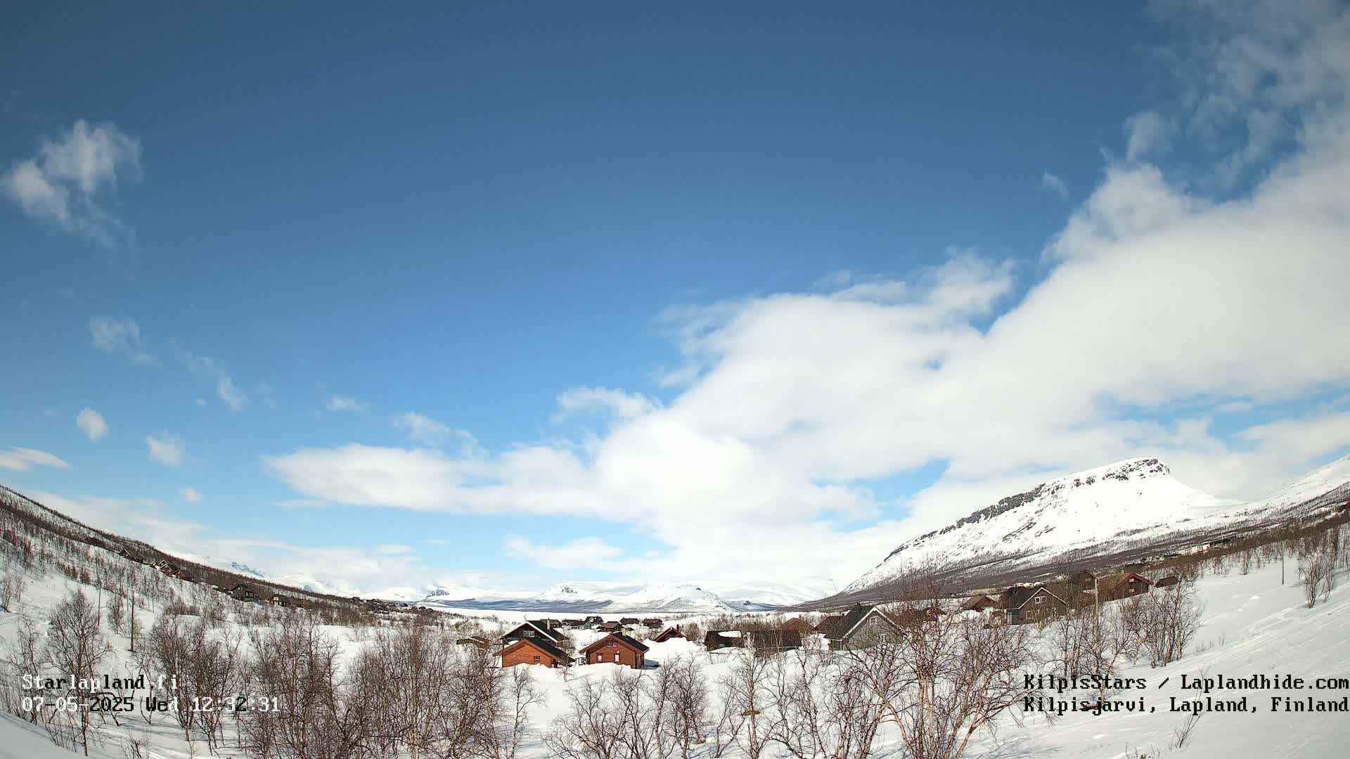A snow-covered valley with a few small buildings nestled amongst leafless trees under a mostly sunny sky with fluffy white clouds.