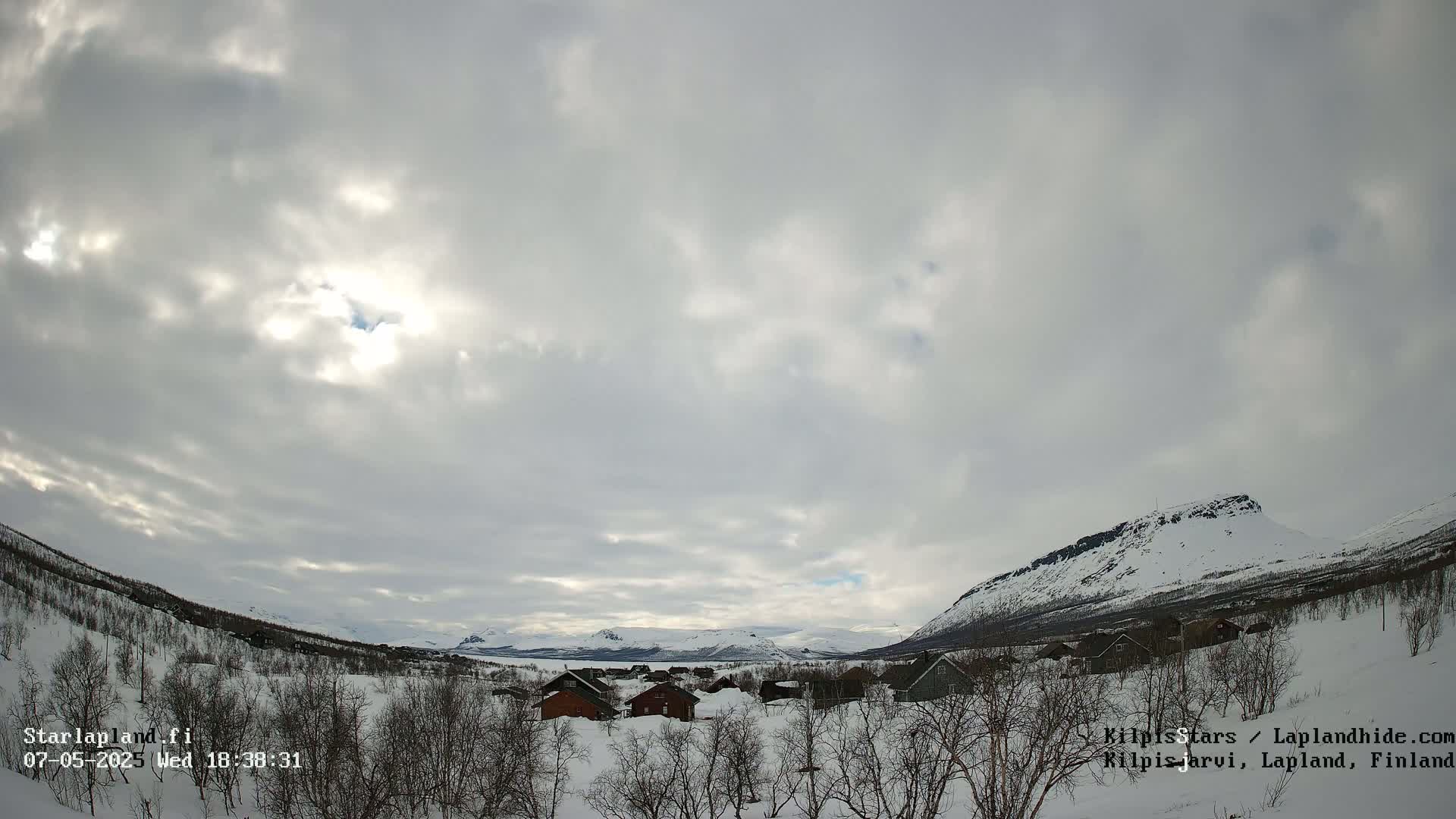 A snowy, mountainous landscape with a small village nestled amongst leafless trees under a mostly cloudy sky, with some sunlight breaking through.