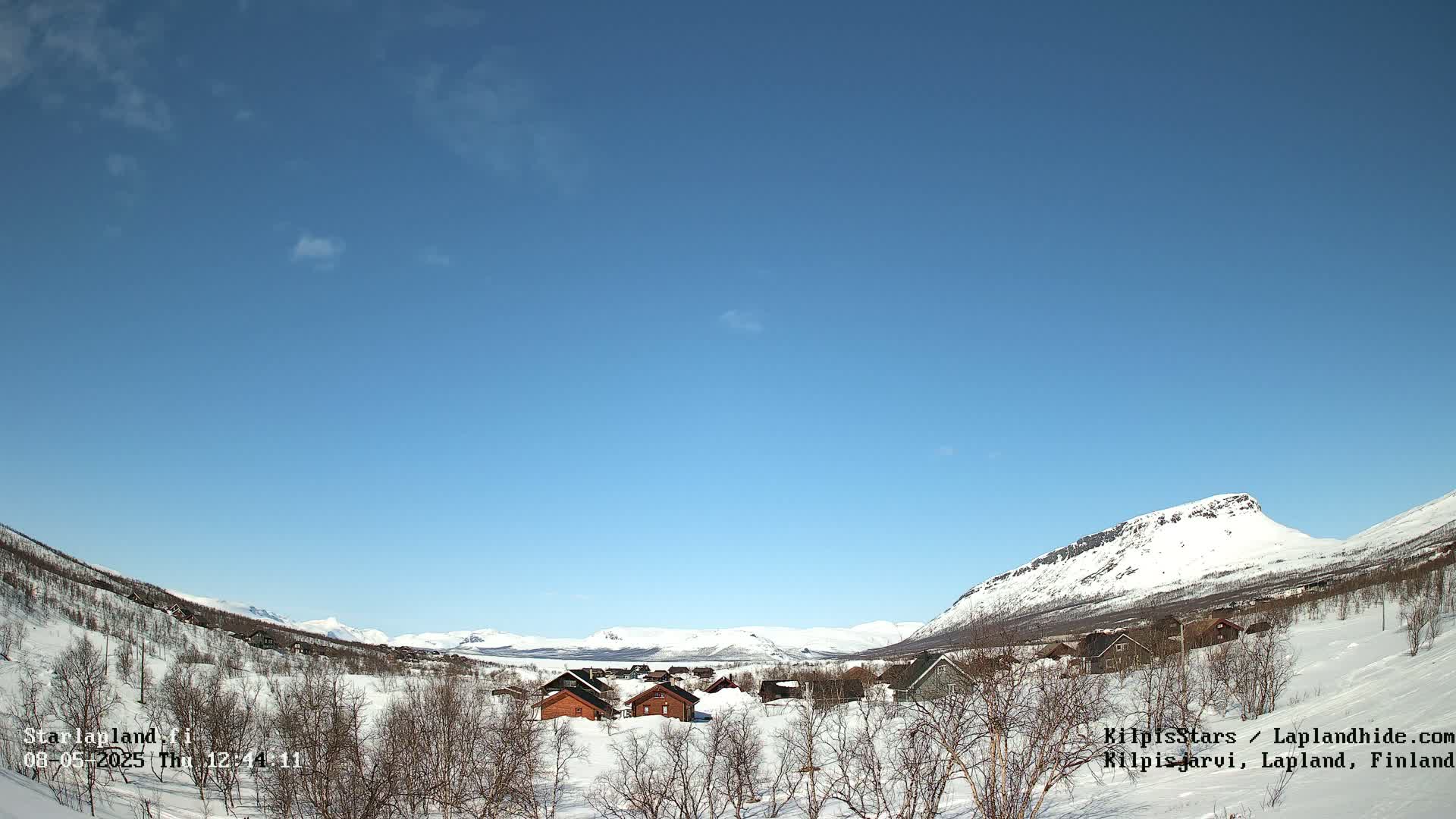 A small village of wooden houses sits in a snowy valley between snow-covered mountains under a clear blue sky.