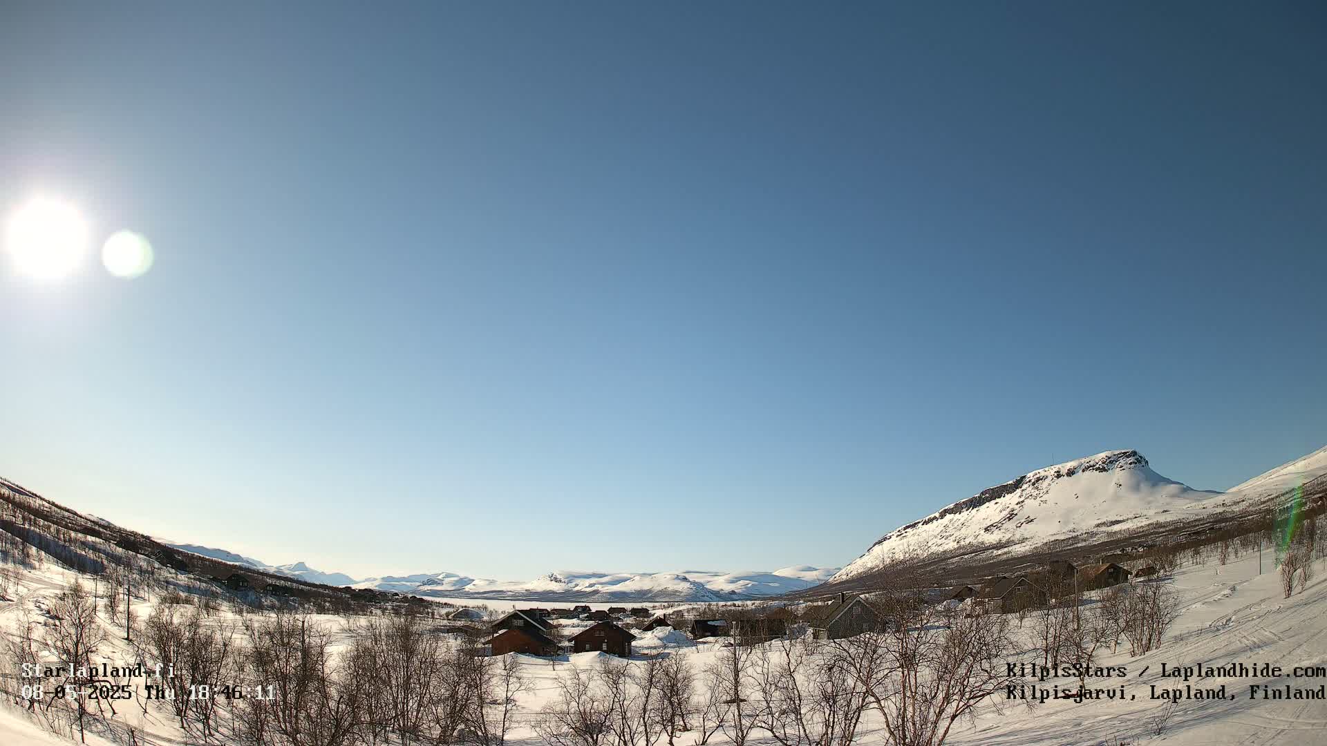 A sunny, snow-covered landscape features a small village nestled among barren trees and snow-capped mountains under a clear blue sky.