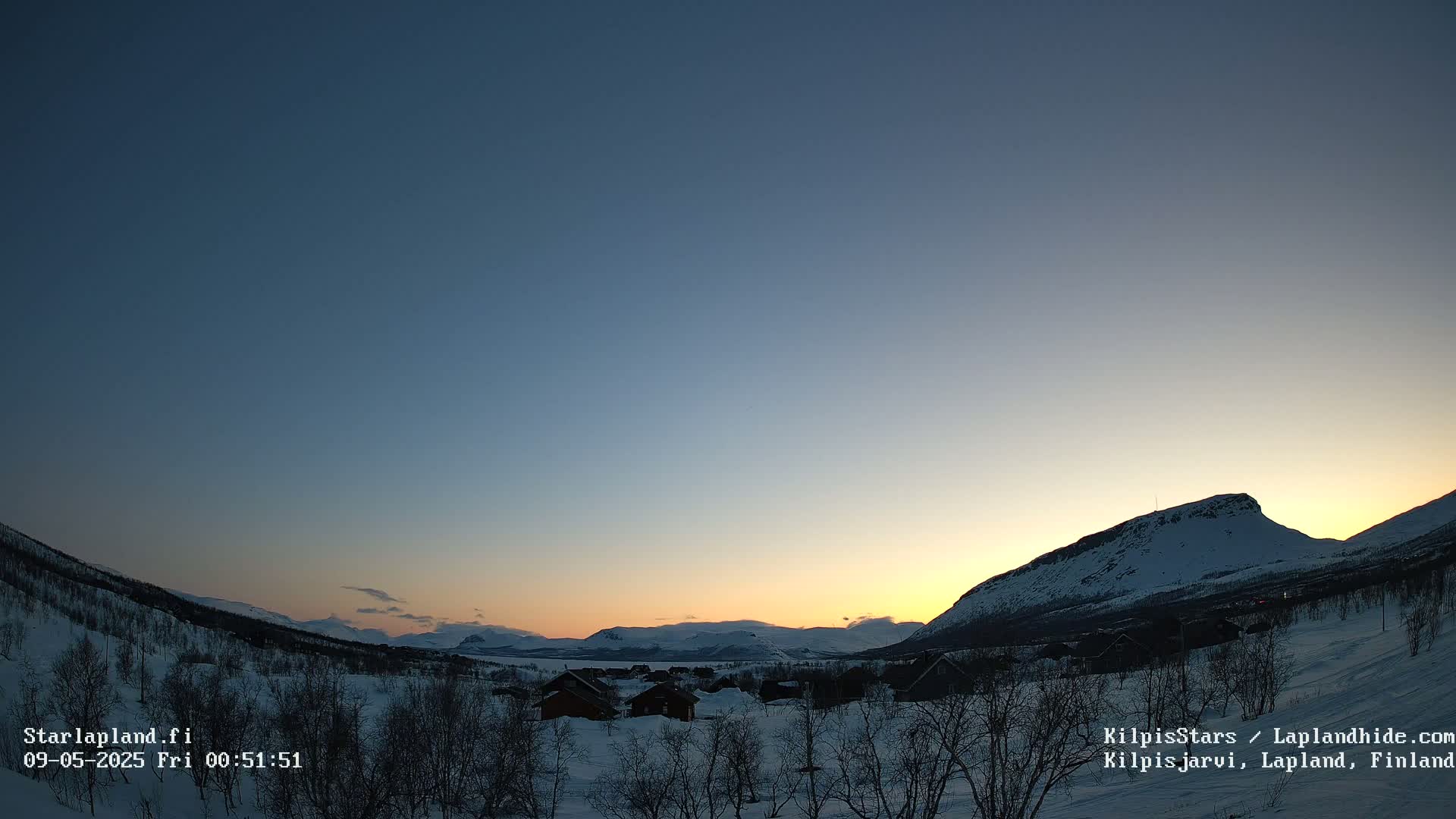 A snow-covered landscape at sunset, with a few houses nestled among bare trees and mountains in the background.