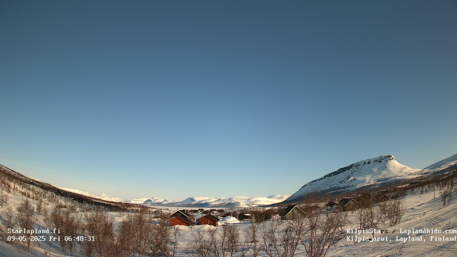 A snow-covered valley with a small cluster of houses nestled amongst leafless trees under a clear blue sky.
