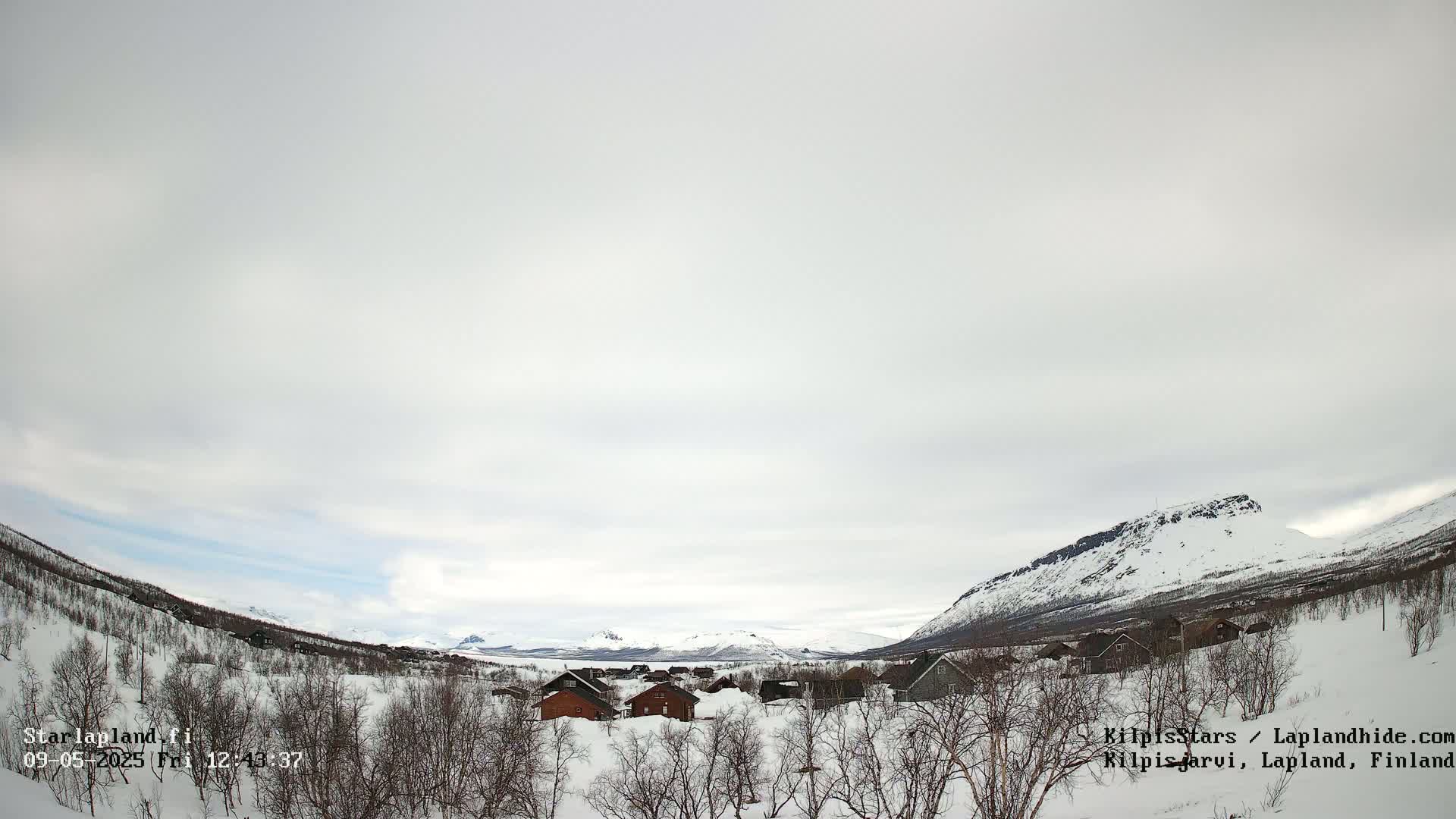 A small snow-covered village nestled in a valley between snow-capped mountains under a mostly cloudy sky.