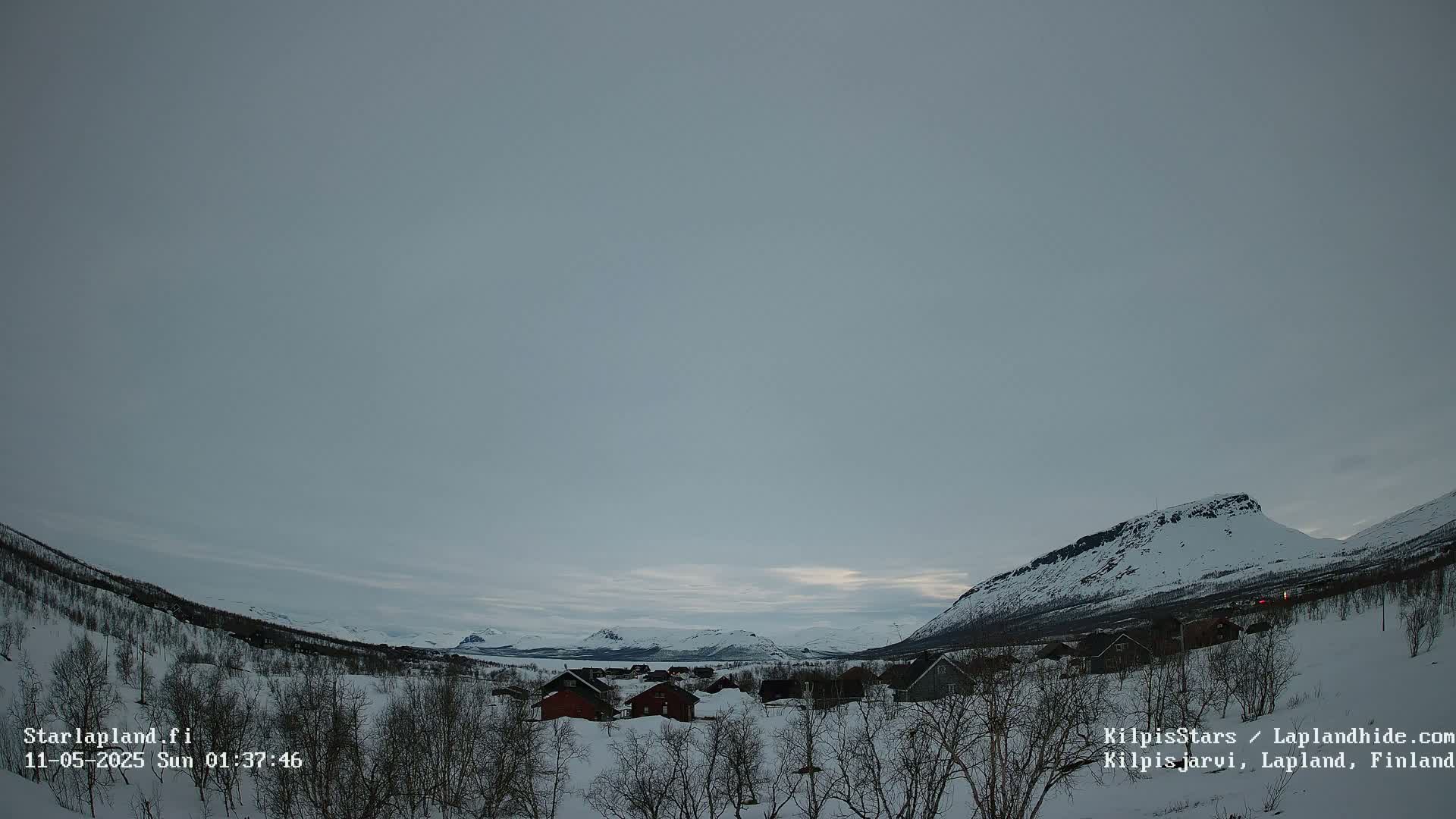 A snow-covered landscape with small houses nestled among leafless trees and mountains in the background under a cloudy, overcast sky.
