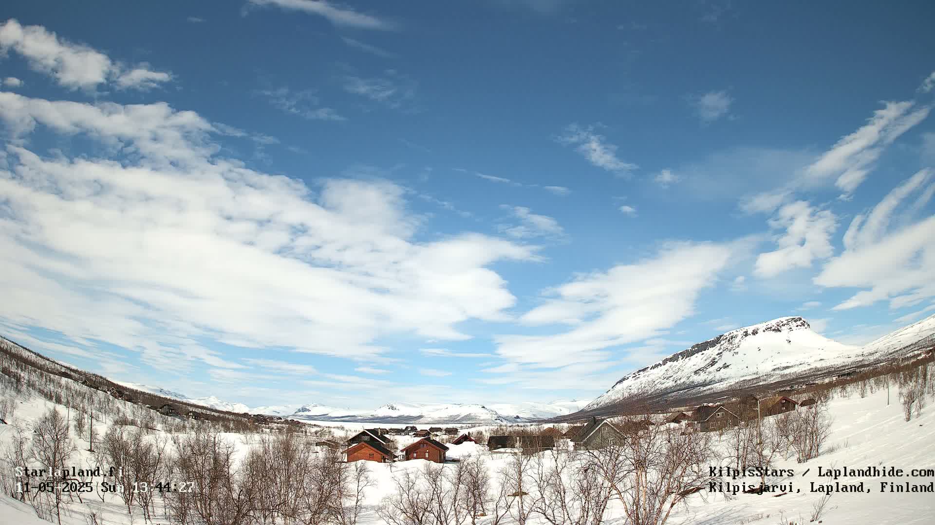 A snow-covered village nestled in a valley between snow-capped mountains under a mostly sunny sky with scattered clouds.