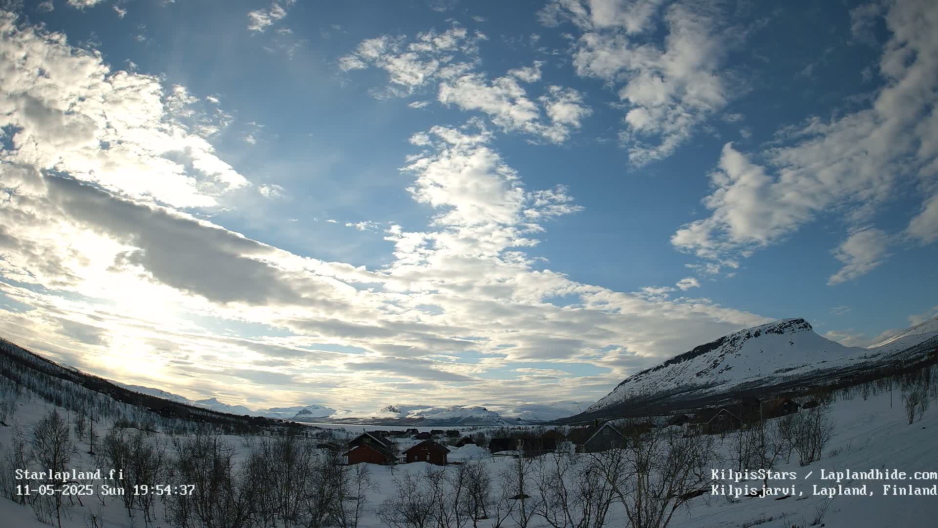 A snow-covered landscape with small houses, sparse trees, and mountains under a partly cloudy, bright sky.