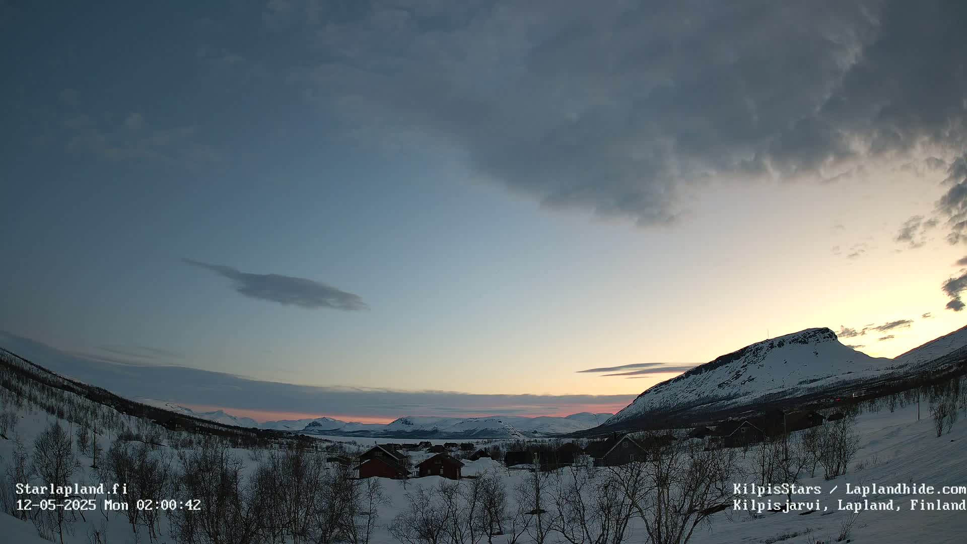 A snow-covered landscape with a few houses and distant mountains is bathed in the soft light of a sunrise or sunset under a partly cloudy sky.