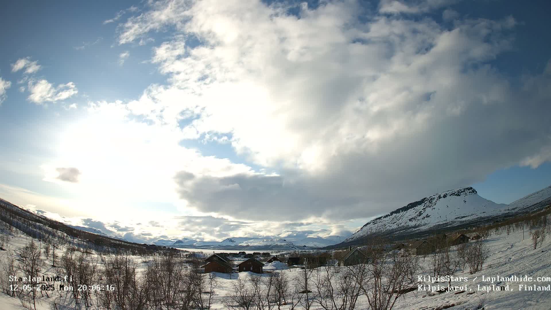 A partly cloudy day reveals a snow-covered landscape with a small village nestled amongst bare trees and mountains in the distance.