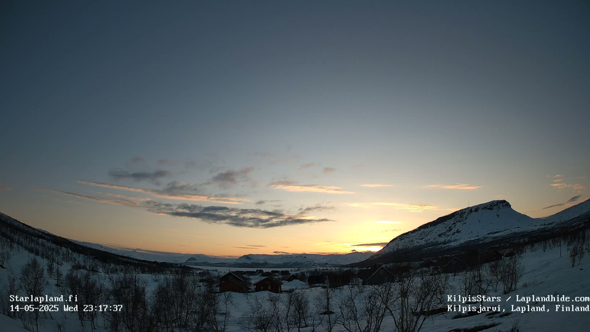 A snowy landscape at sunset, with a few buildings nestled among bare trees in the foreground and mountains in the background under a partly cloudy sky.