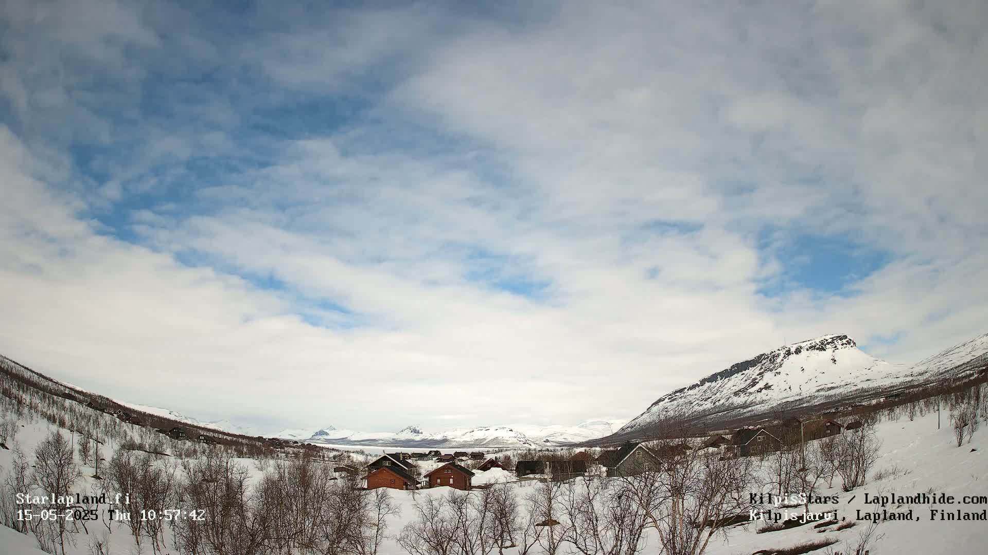 A snow-covered village nestled in a valley between snow-capped mountains under a partly cloudy sky.