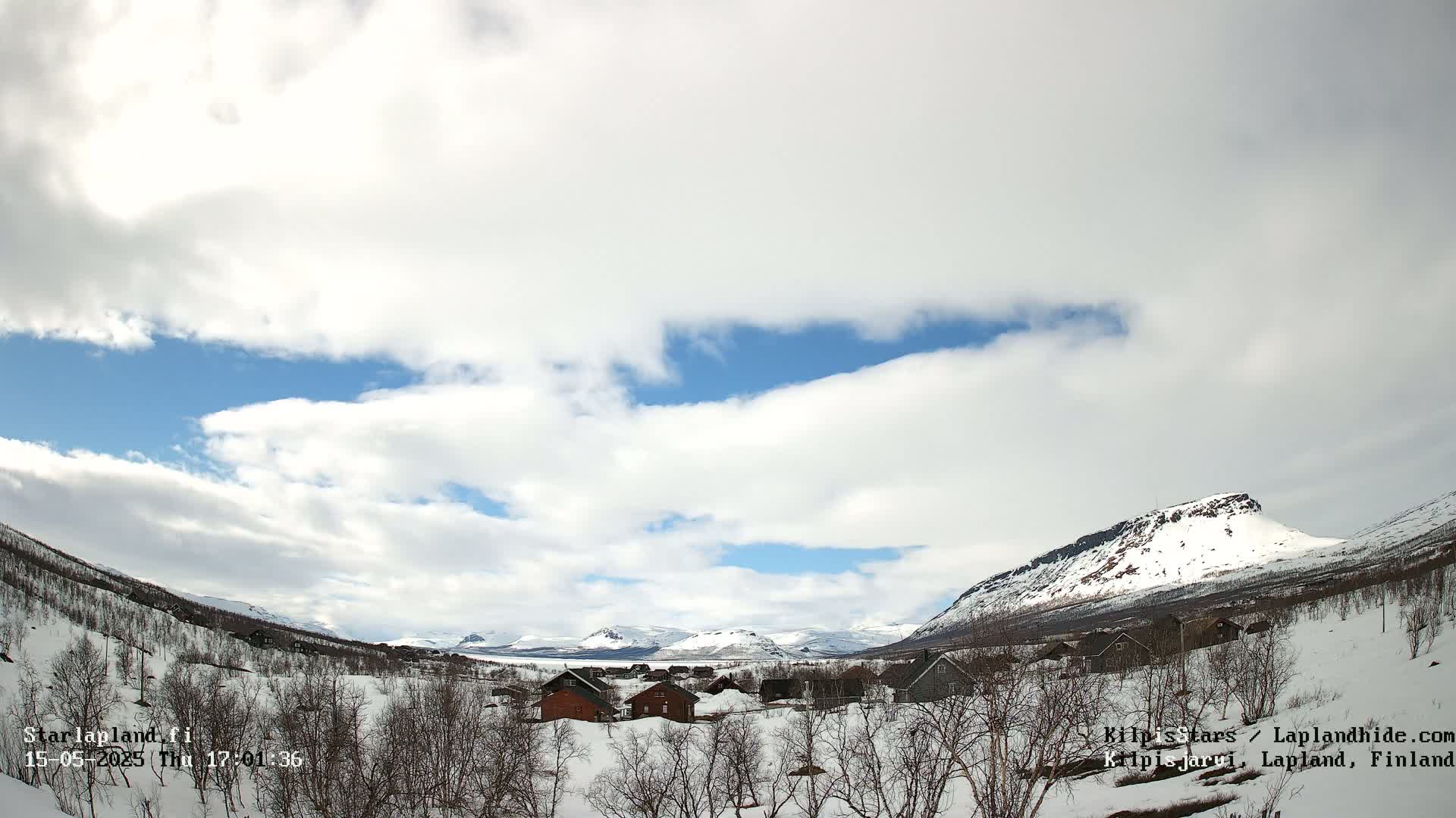 A partly cloudy sky over a snow-covered valley with small buildings and mountains in the background.