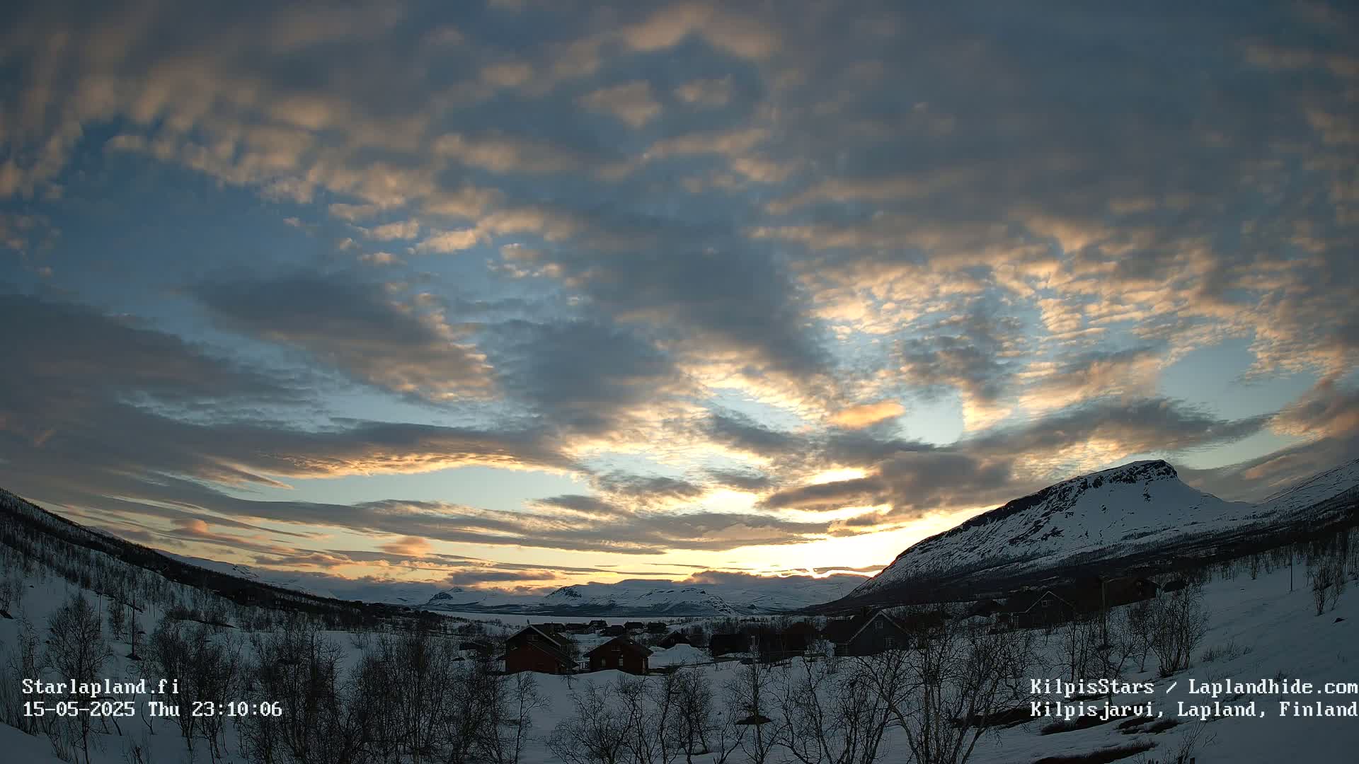 A snow-covered landscape at sunset, with a cloudy sky displaying various shades of orange, pink, and blue,  and mountains in the background.