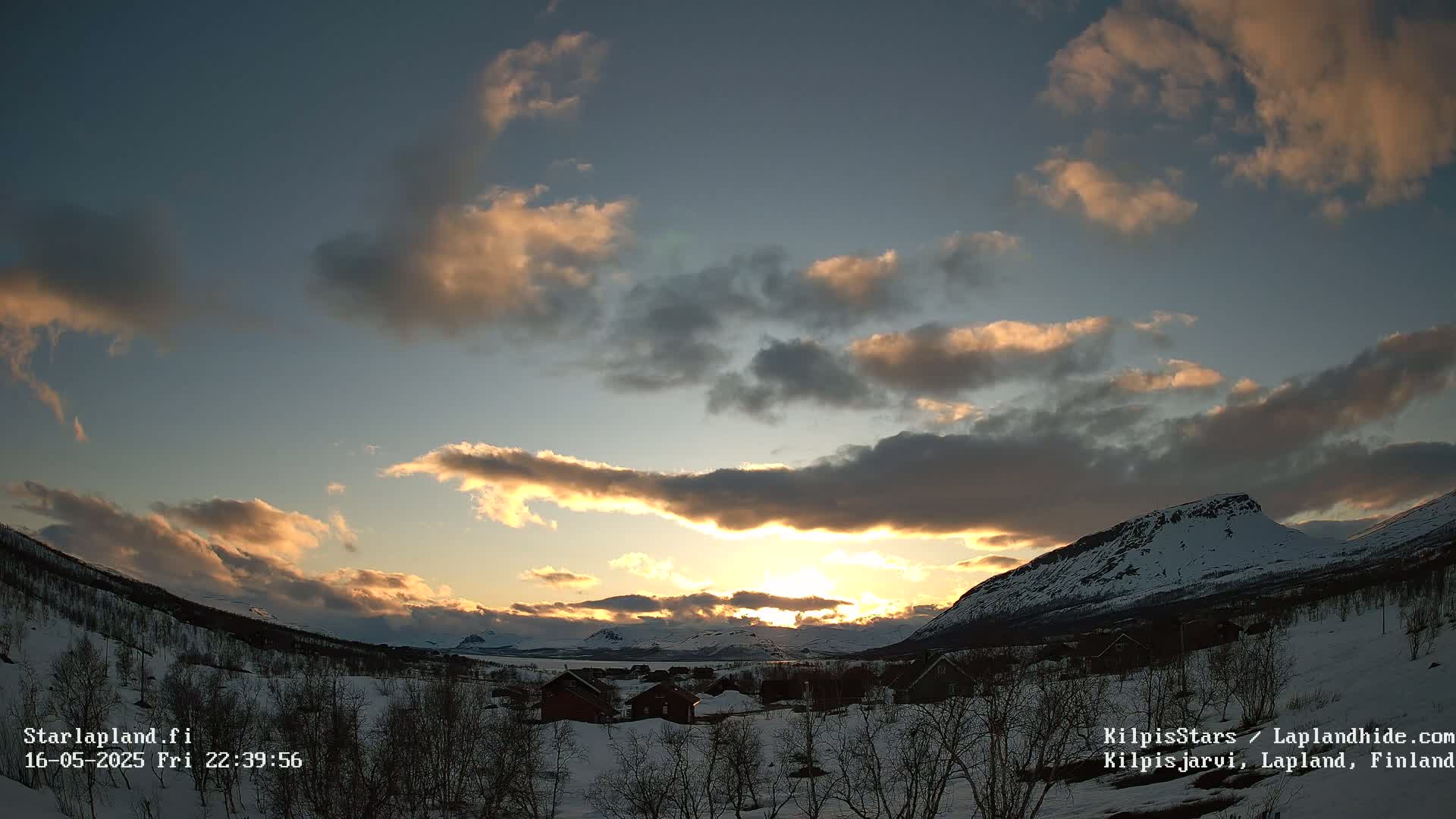 A snowy, mountainous landscape is bathed in the warm light of a setting sun, partially obscured by fluffy clouds.