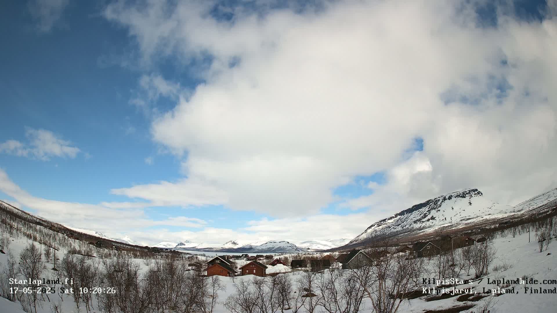 A partly cloudy sky over a snow-covered village nestled in a valley between snow-capped mountains.