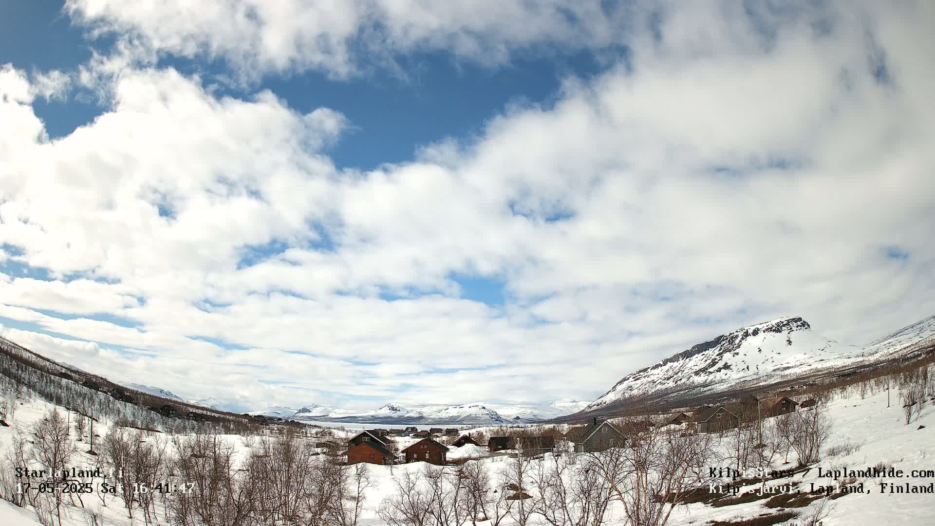 A snow-covered village nestled in a valley between snow-capped mountains under a partly cloudy sky.