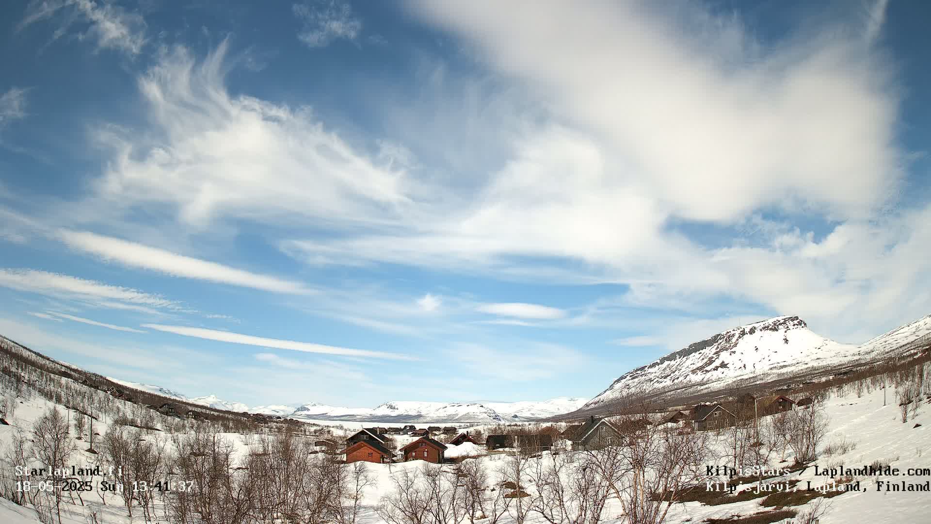 A snow-covered village nestled in a valley between snow-capped mountains under a partly cloudy blue sky.
