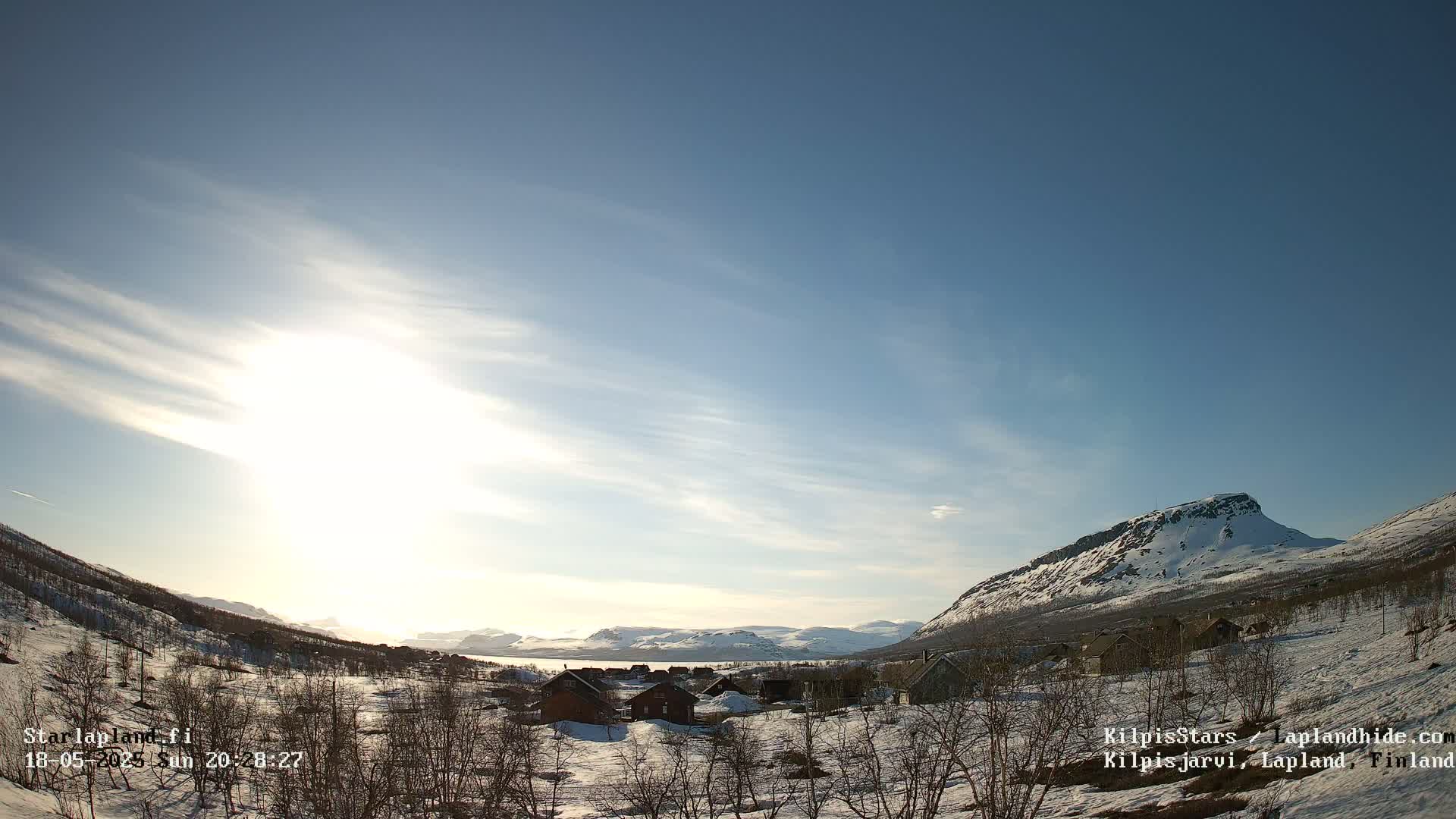 A sunny, partly cloudy day reveals a snow-covered valley with a few houses nestled amongst leafless trees and mountains in the background.