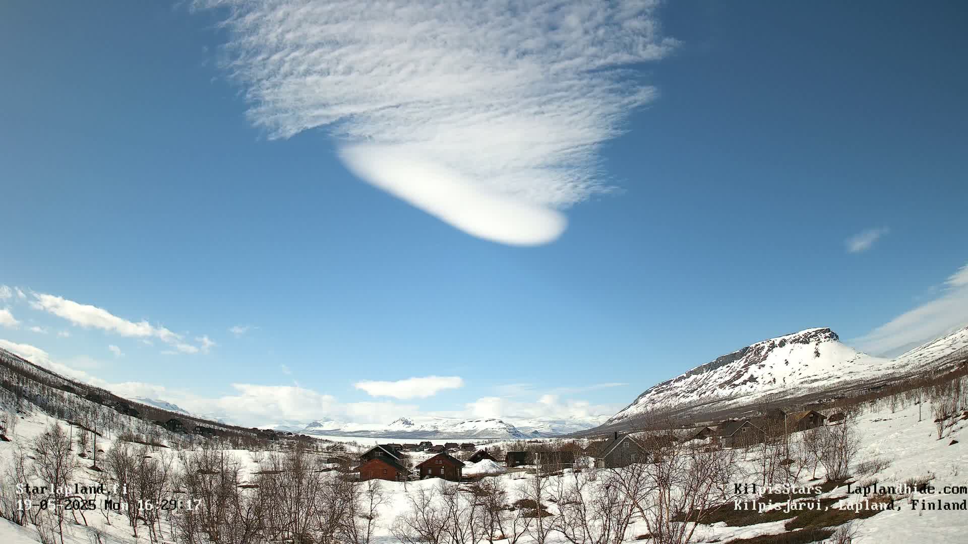 A mostly clear blue sky with a lenticular cloud formation above a snow-covered valley with small houses and distant mountains.