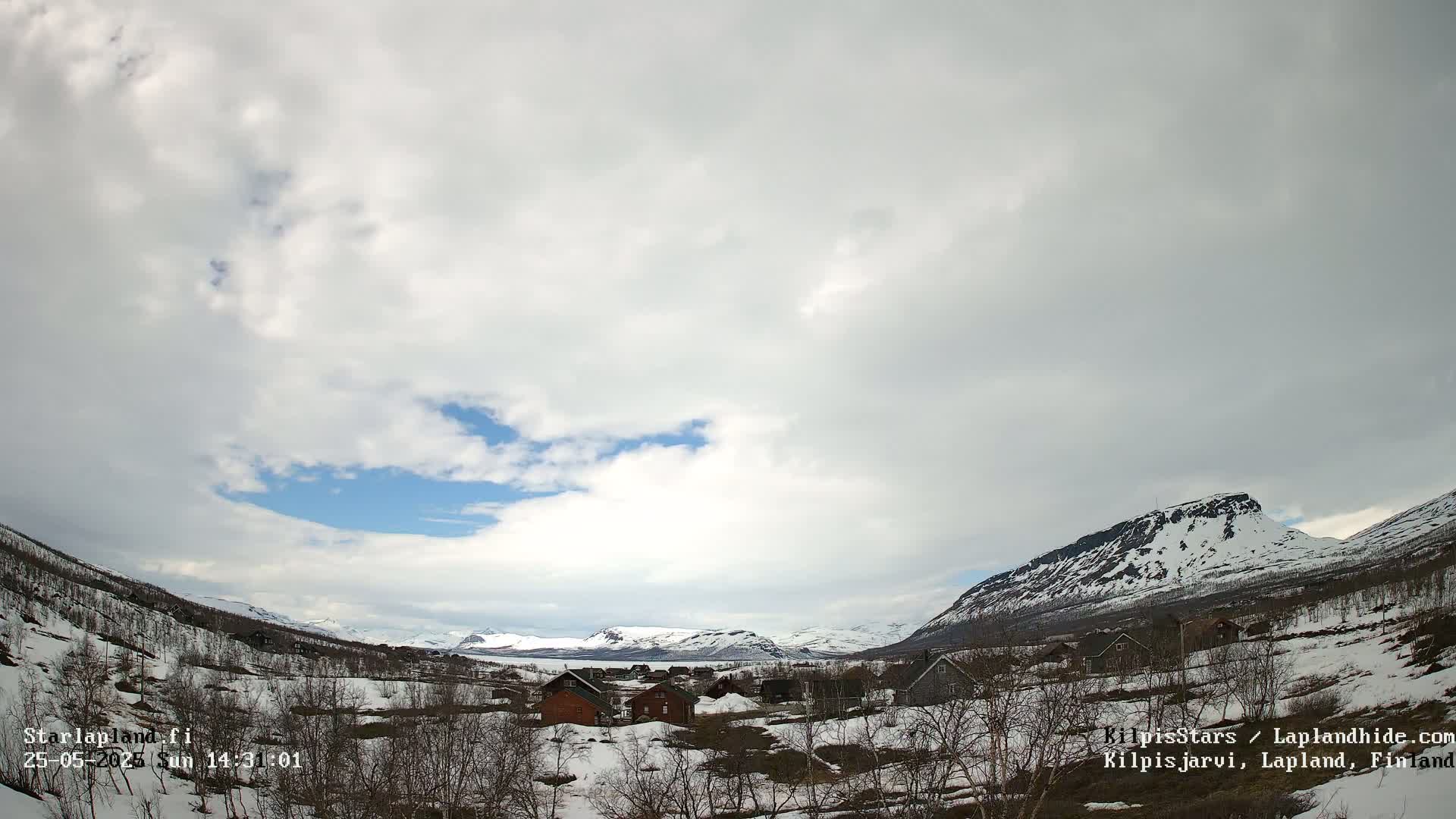 A partly cloudy sky overlooks a snow-dusted village nestled in a valley between mountains.