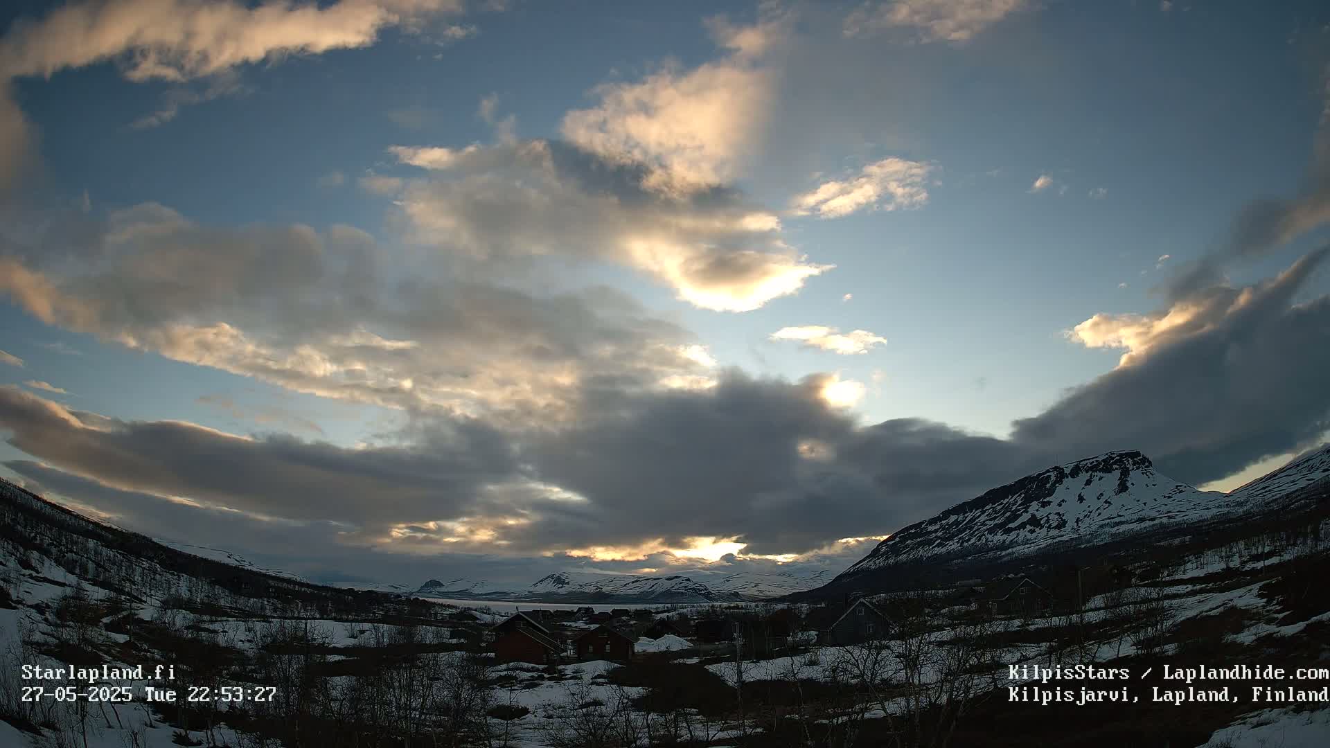A partially cloudy sunset illuminates a snow-covered valley with small houses and mountains in the background.