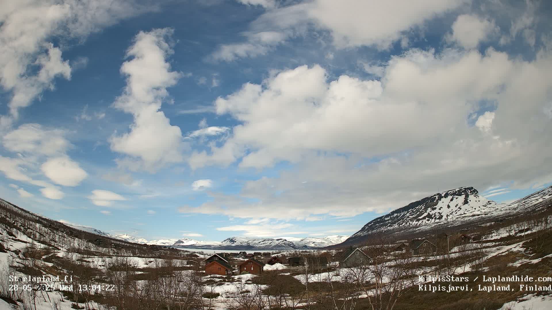 A mostly sunny day with scattered clouds over a snow-dusted valley, featuring a small village nestled amongst low hills and mountains in the distance.