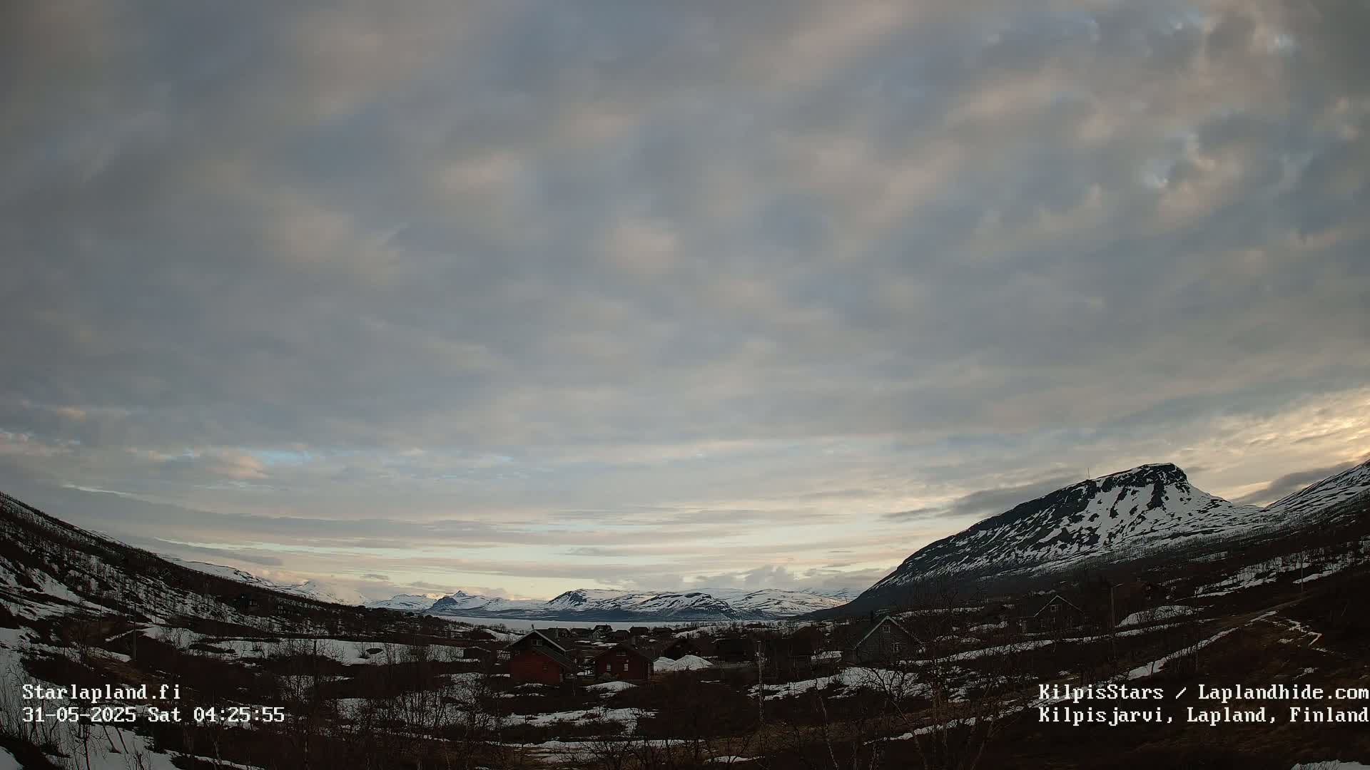 A small village nestled in a valley between snow-capped mountains under a mostly cloudy sky.