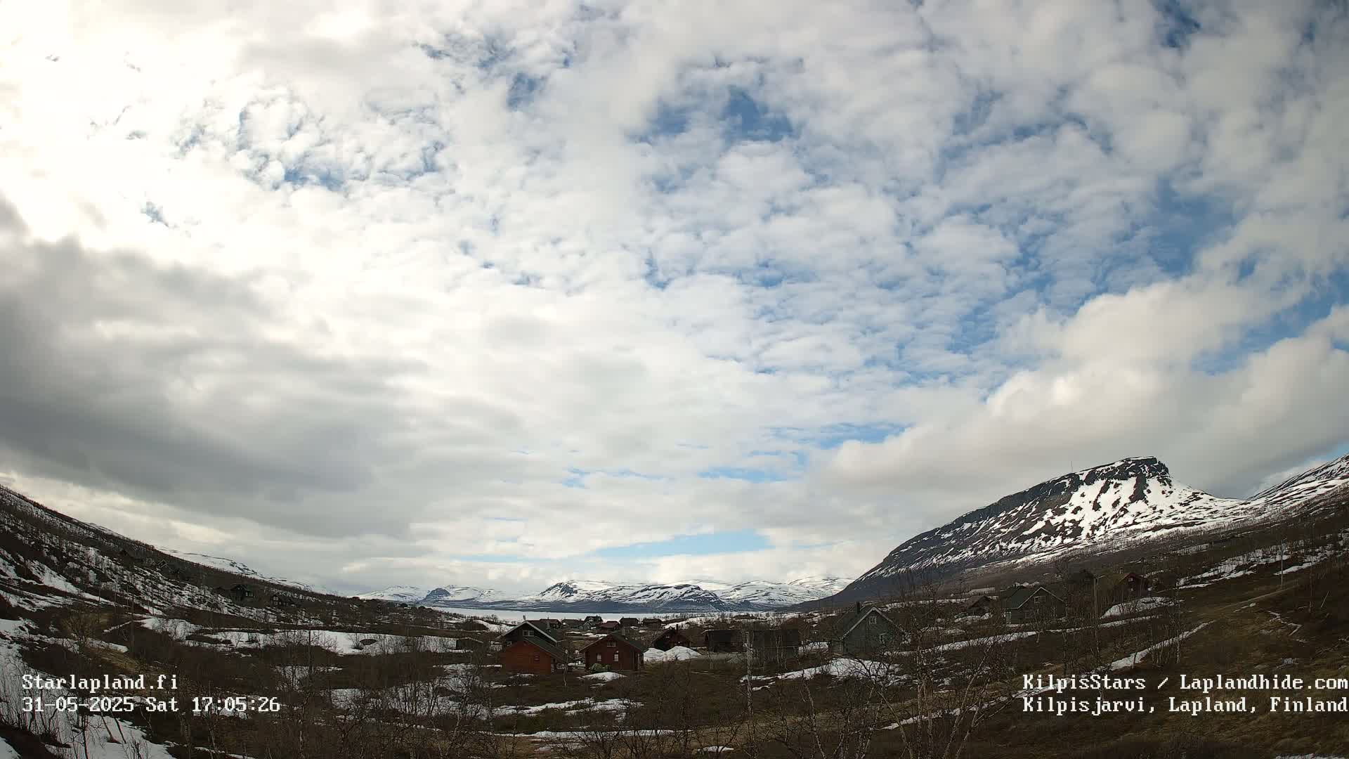 A partly cloudy sky over a small village nestled in a valley between snow-capped mountains.
