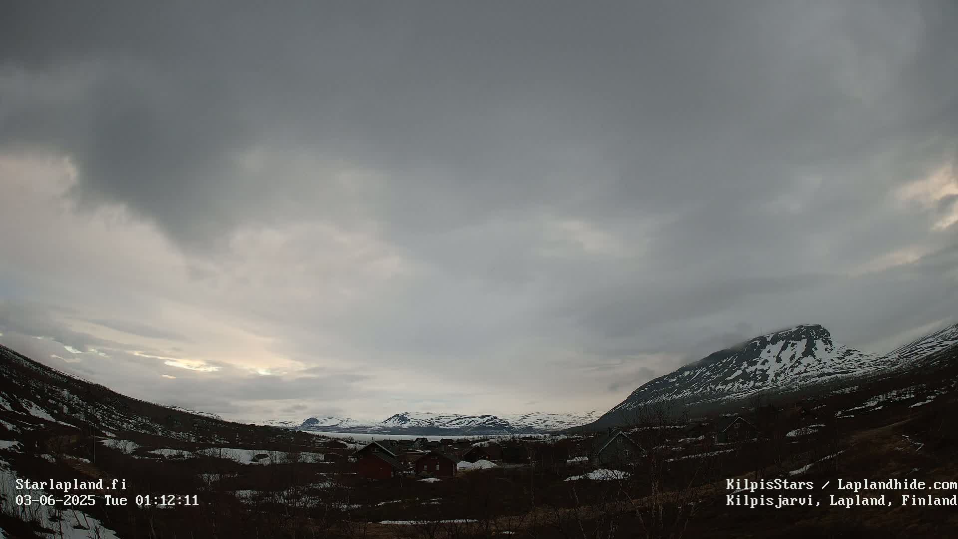 A small village nestled in a valley between snow-capped mountains under a mostly cloudy sky.