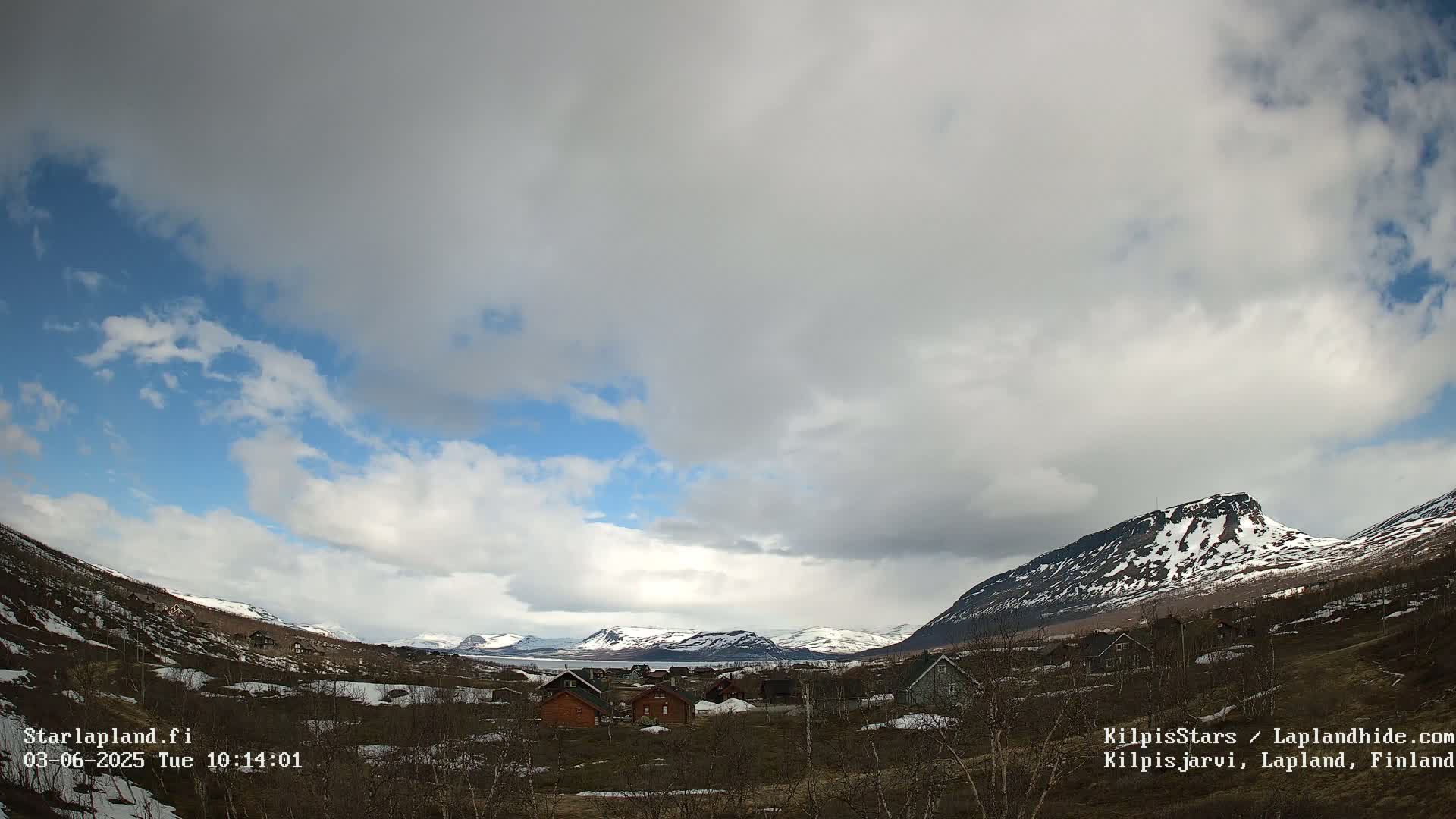 A partly cloudy sky hangs over a small village nestled in a valley between snow-capped mountains.