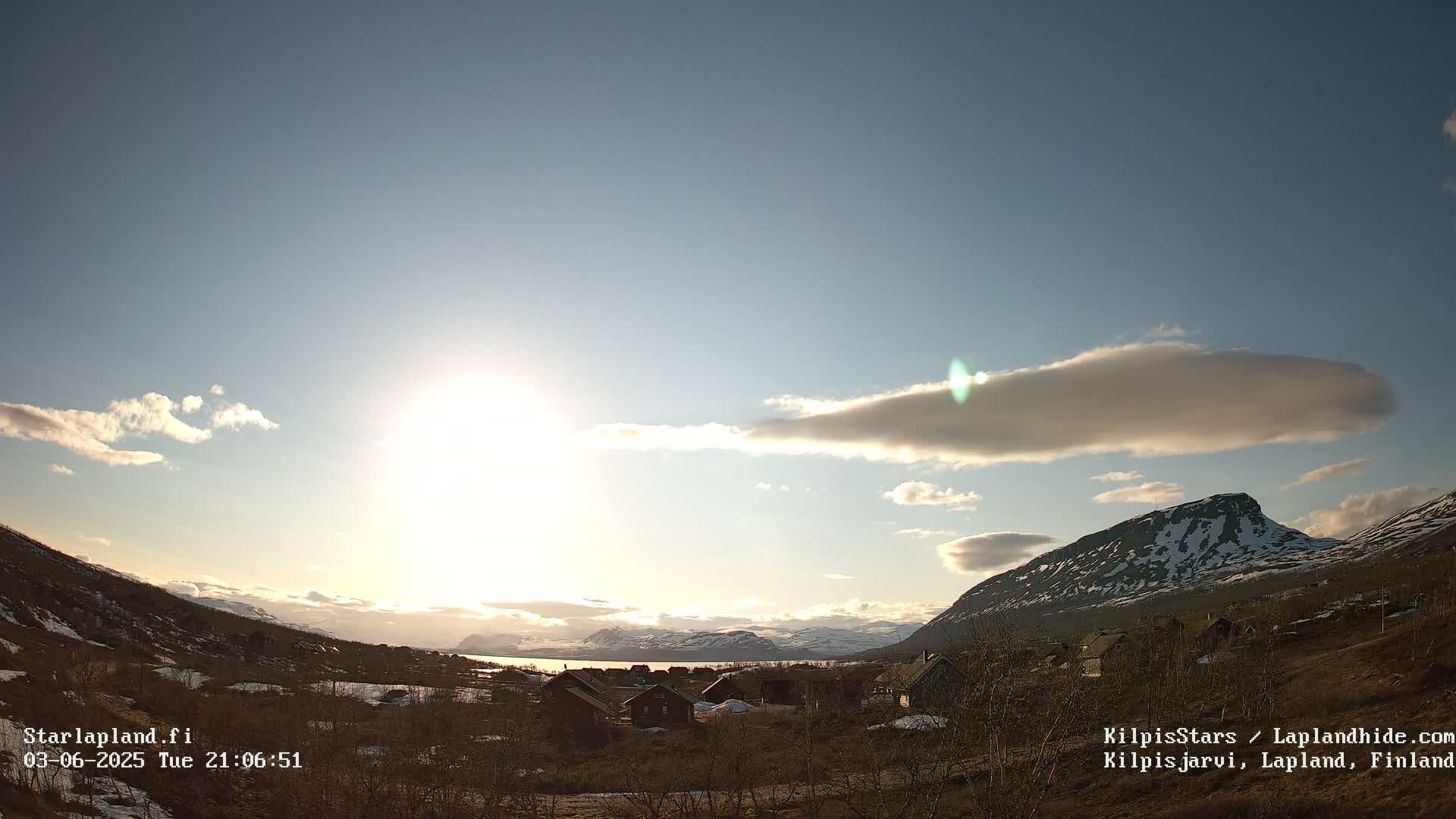 A partly sunny sky over a small village nestled in a valley between snow-capped mountains.