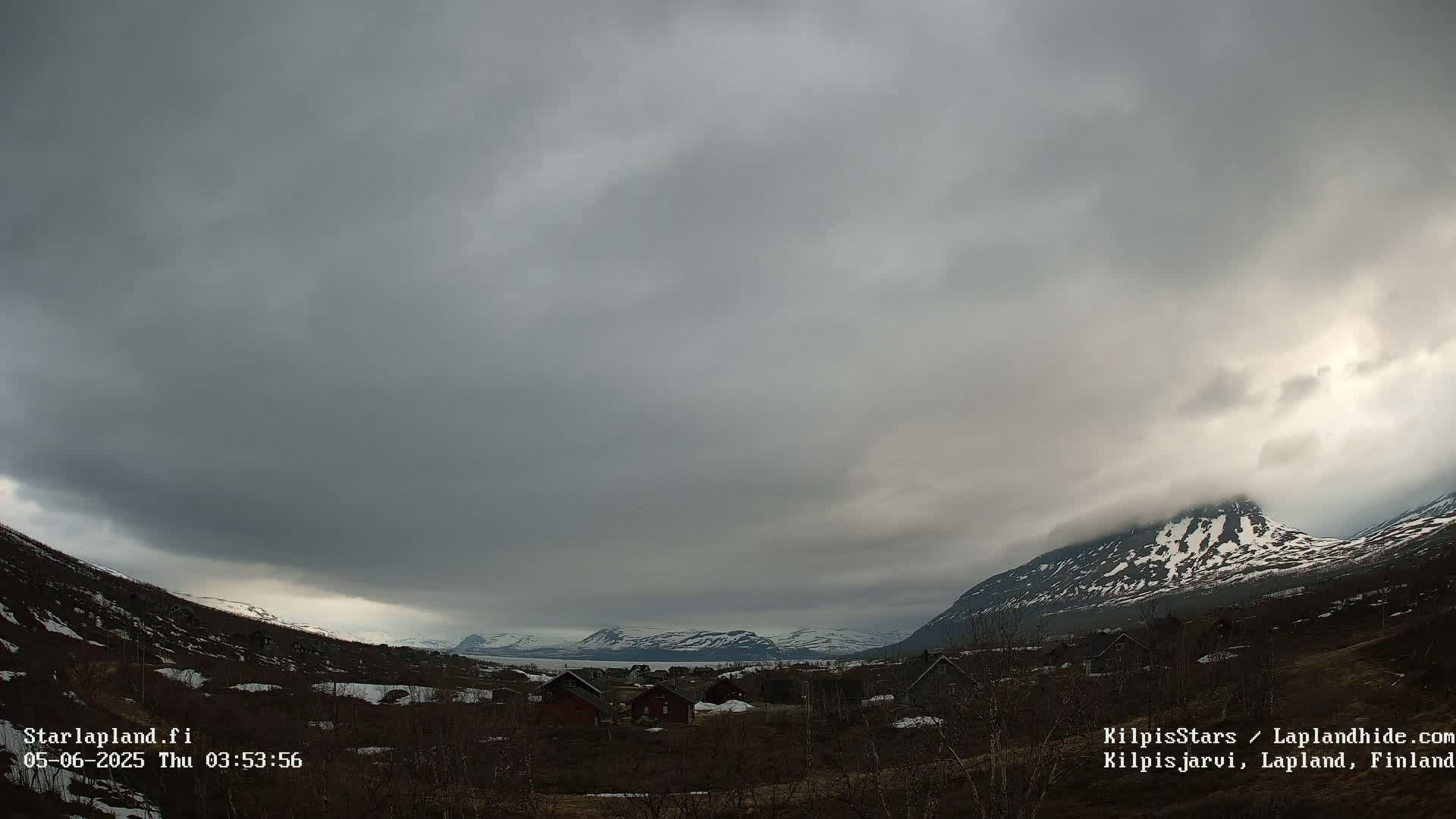 A valley with a few houses is nestled between snow-capped mountains under an overcast sky.