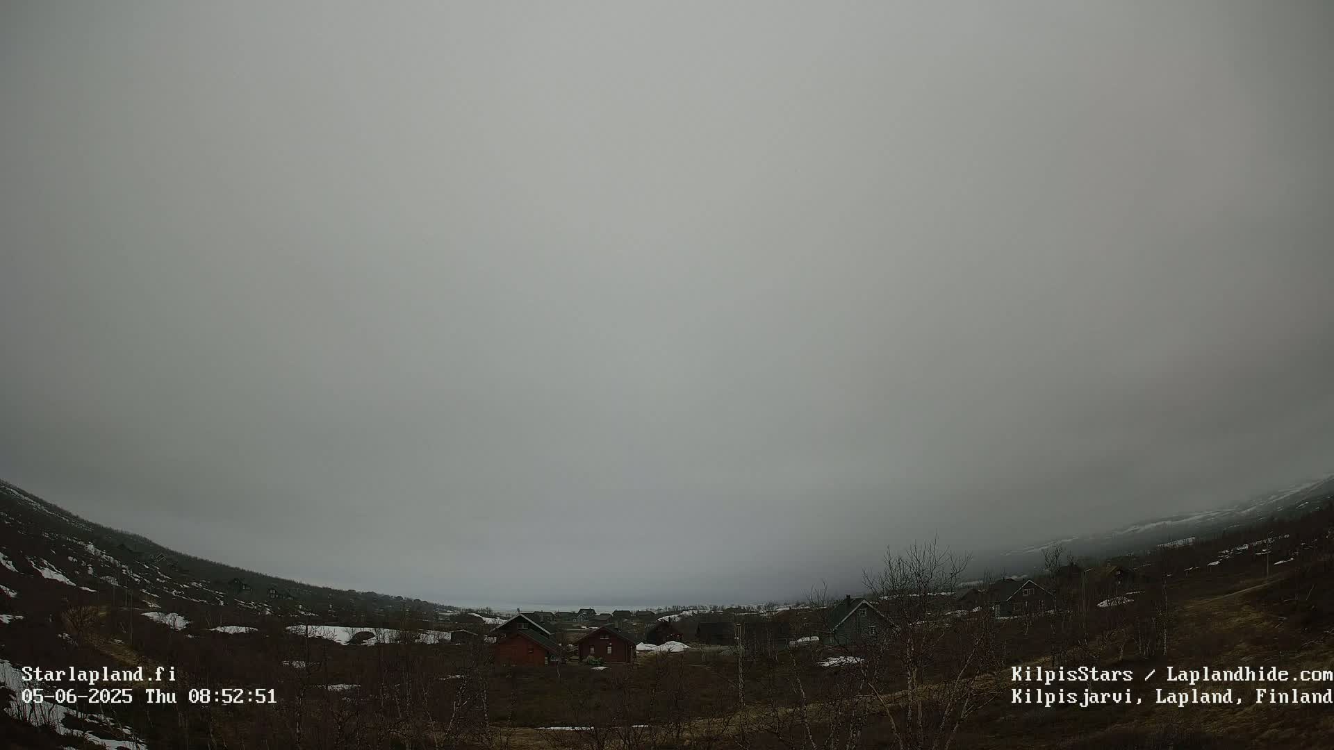 A foggy, overcast day reveals a sparsely populated landscape with small houses nestled in a valley, surrounded by low-lying vegetation and patches of snow.