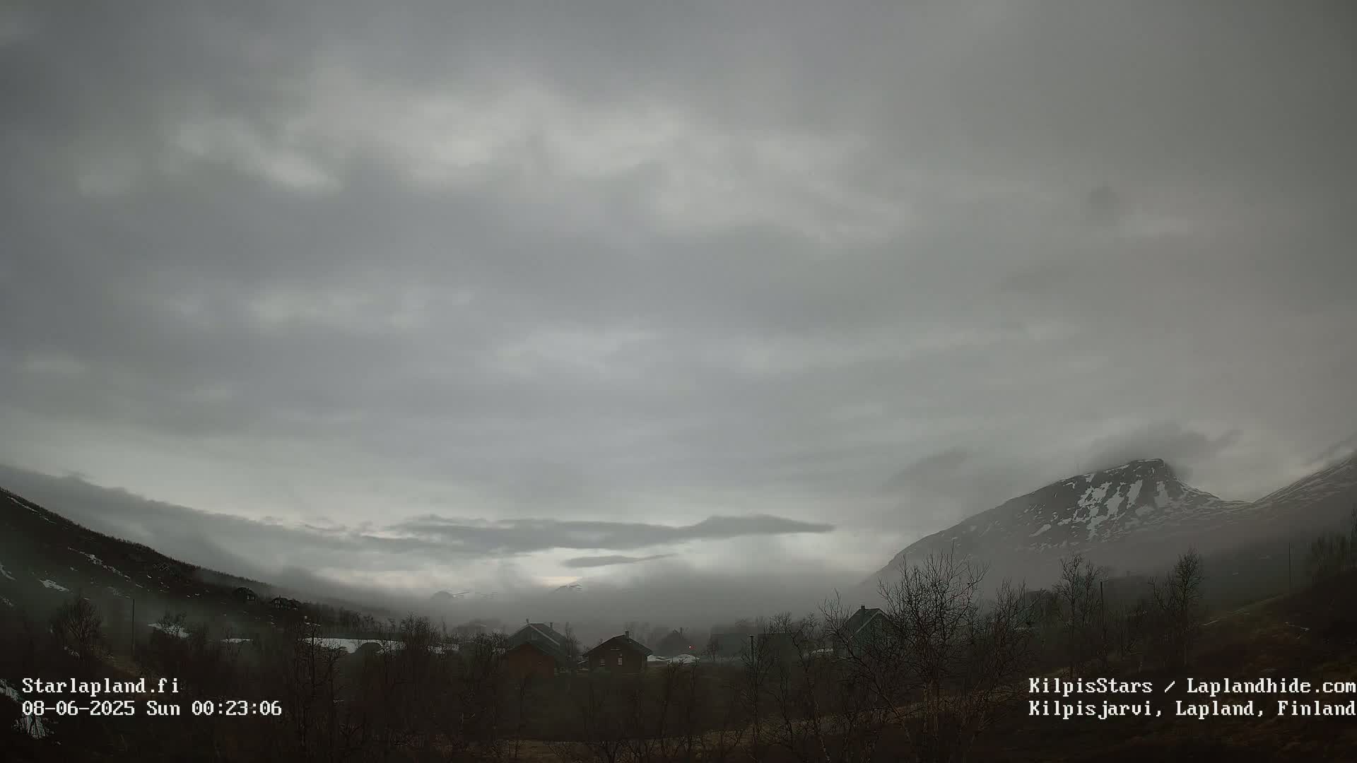 A valley nestled between snow-capped mountains is shrouded in a thick, gray fog under an overcast sky.