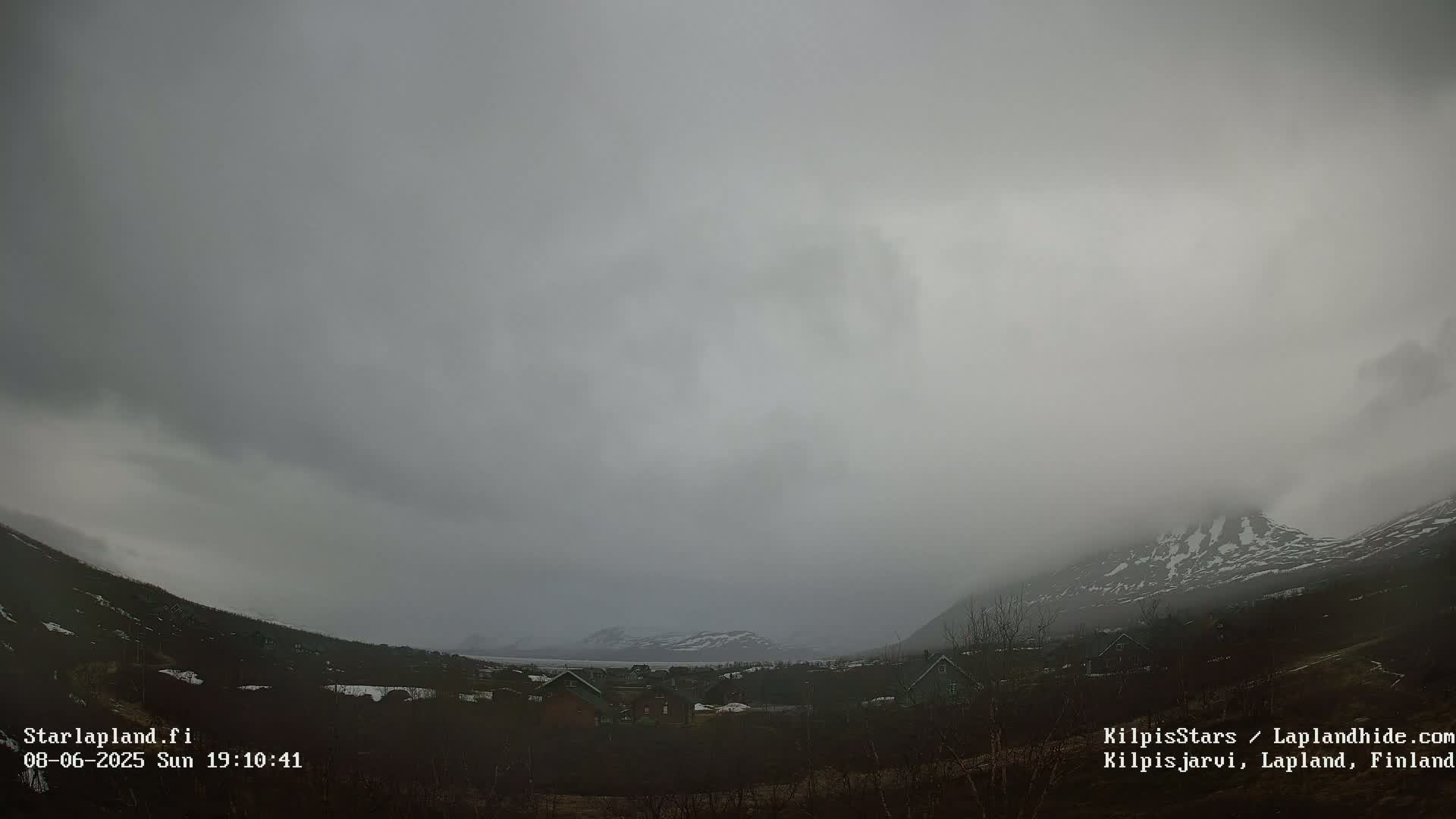 A foggy, overcast day reveals a small village nestled in a valley between snow-capped mountains.