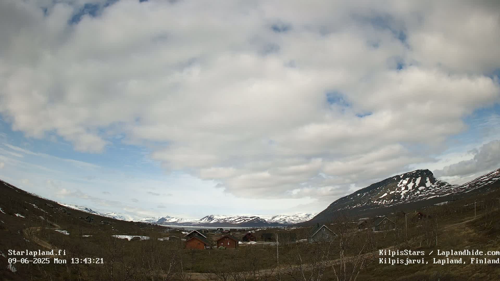 A mostly cloudy sky overlooks a small village nestled in a valley between snow-capped mountains.