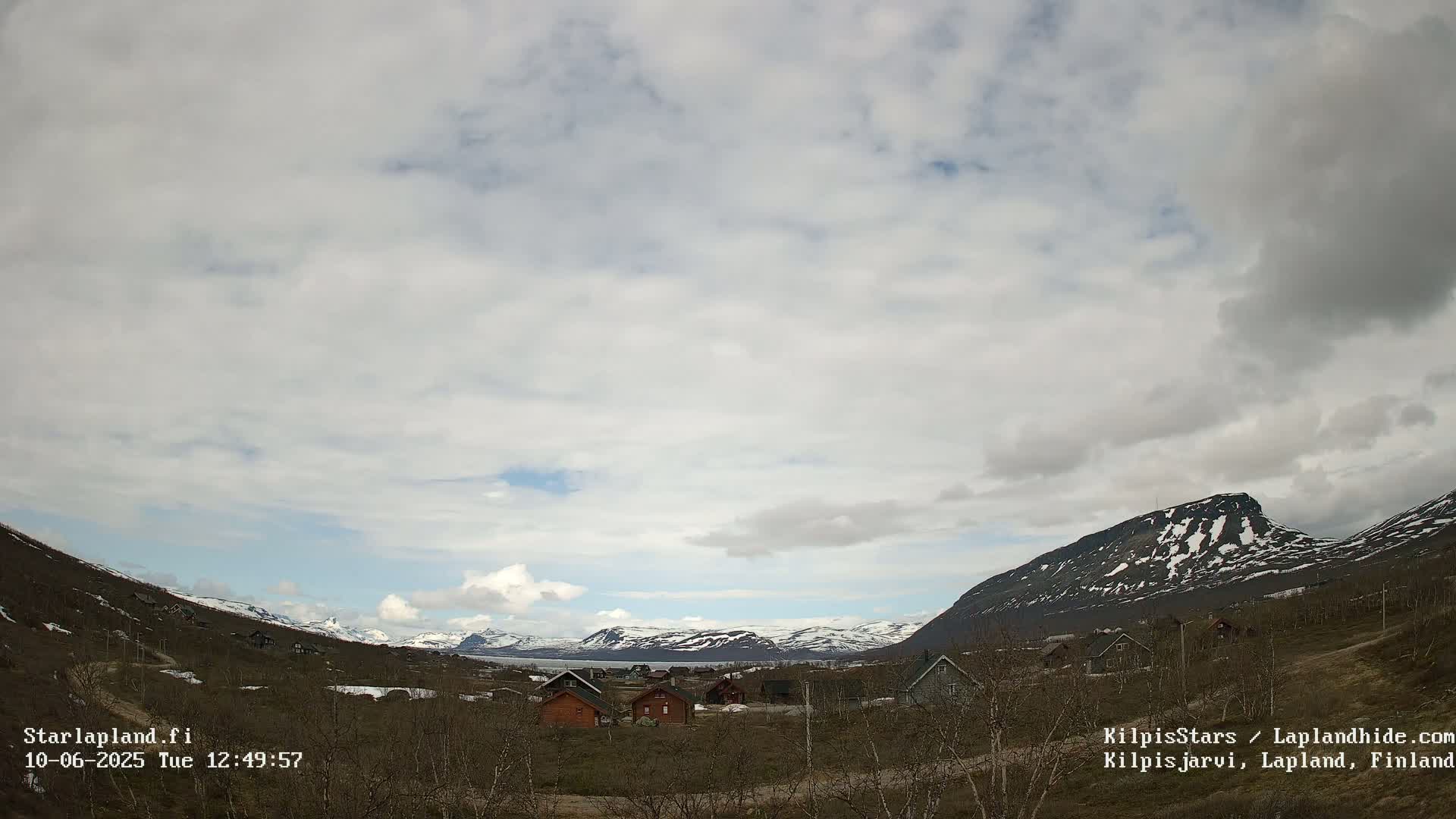 A small village of cabins nestled in a valley between snow-capped mountains under a partly cloudy sky.