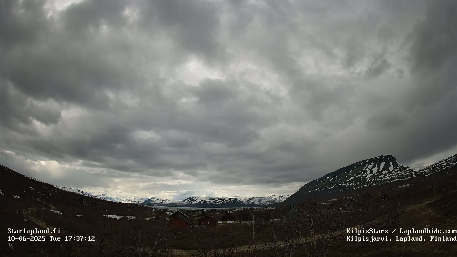 A cloudy day reveals a valley with a small cluster of houses nestled between snow-dusted mountains.