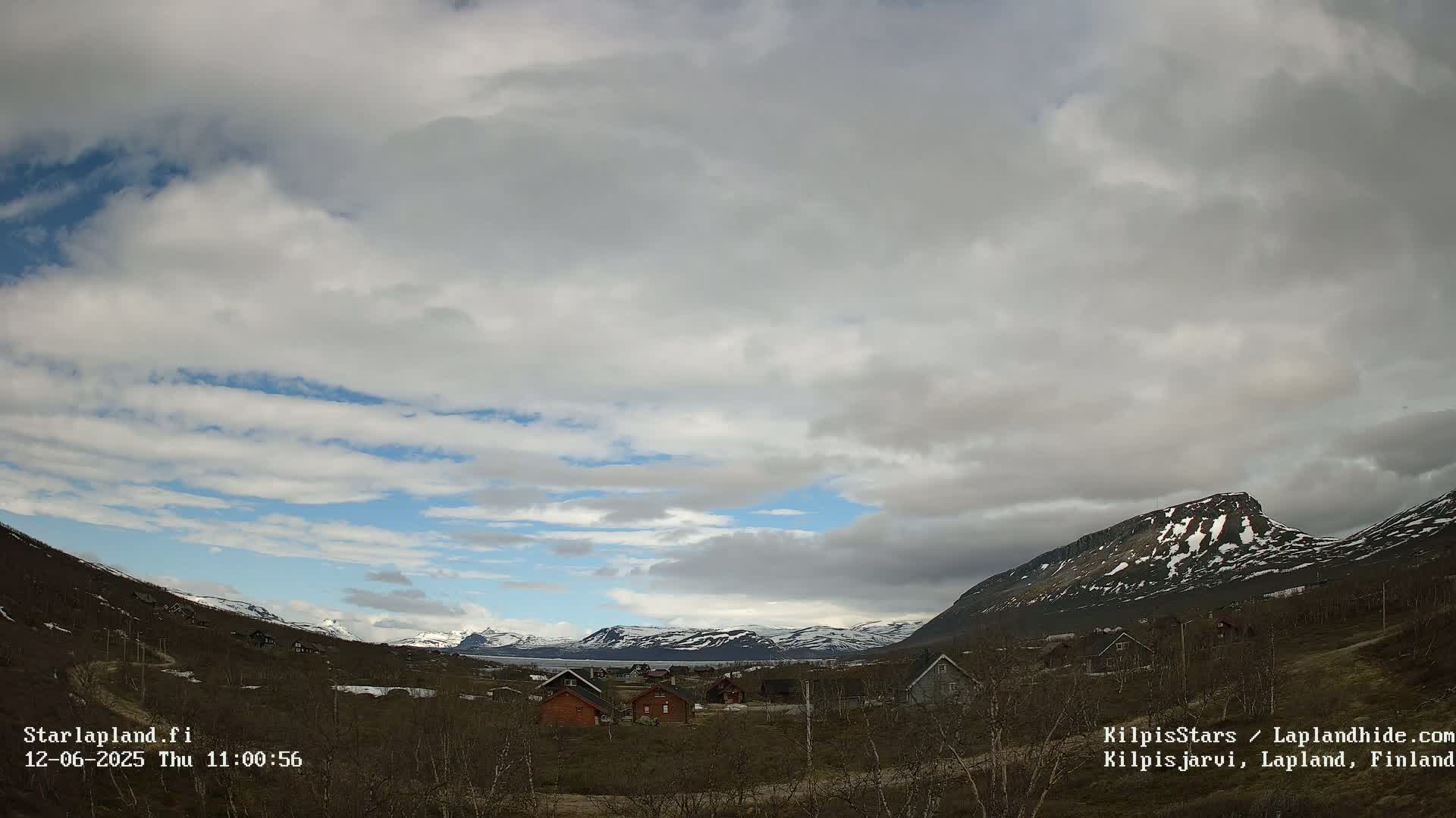 A small village nestled in a valley between snow-capped mountains under a partly cloudy sky.